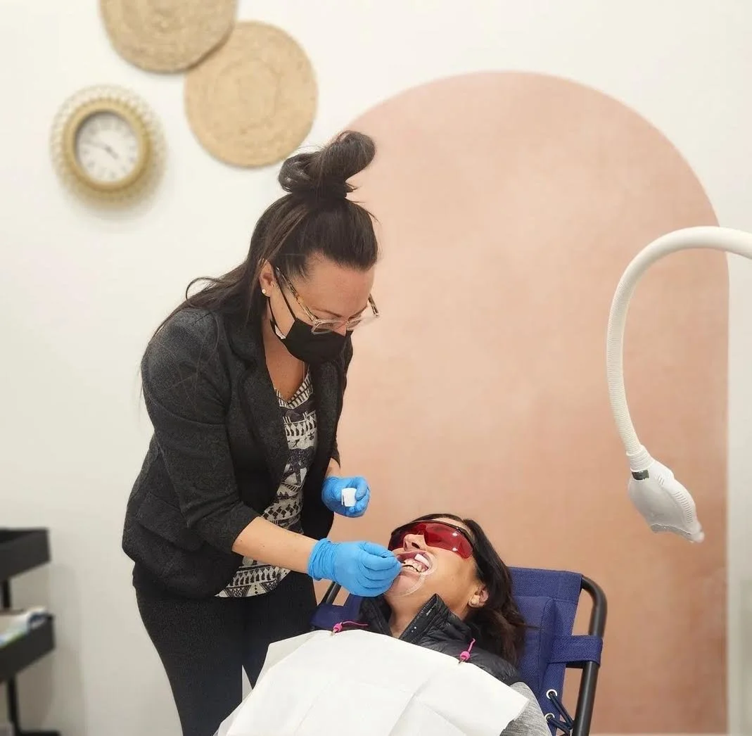 A dental clinic scene where a dental hygienist is performing teeth cleaning on a female patient who is lying in a dental chair. The patient is wearing protective glasses, and the hygienist is wearing a black mask and blue gloves, holding dental tools.