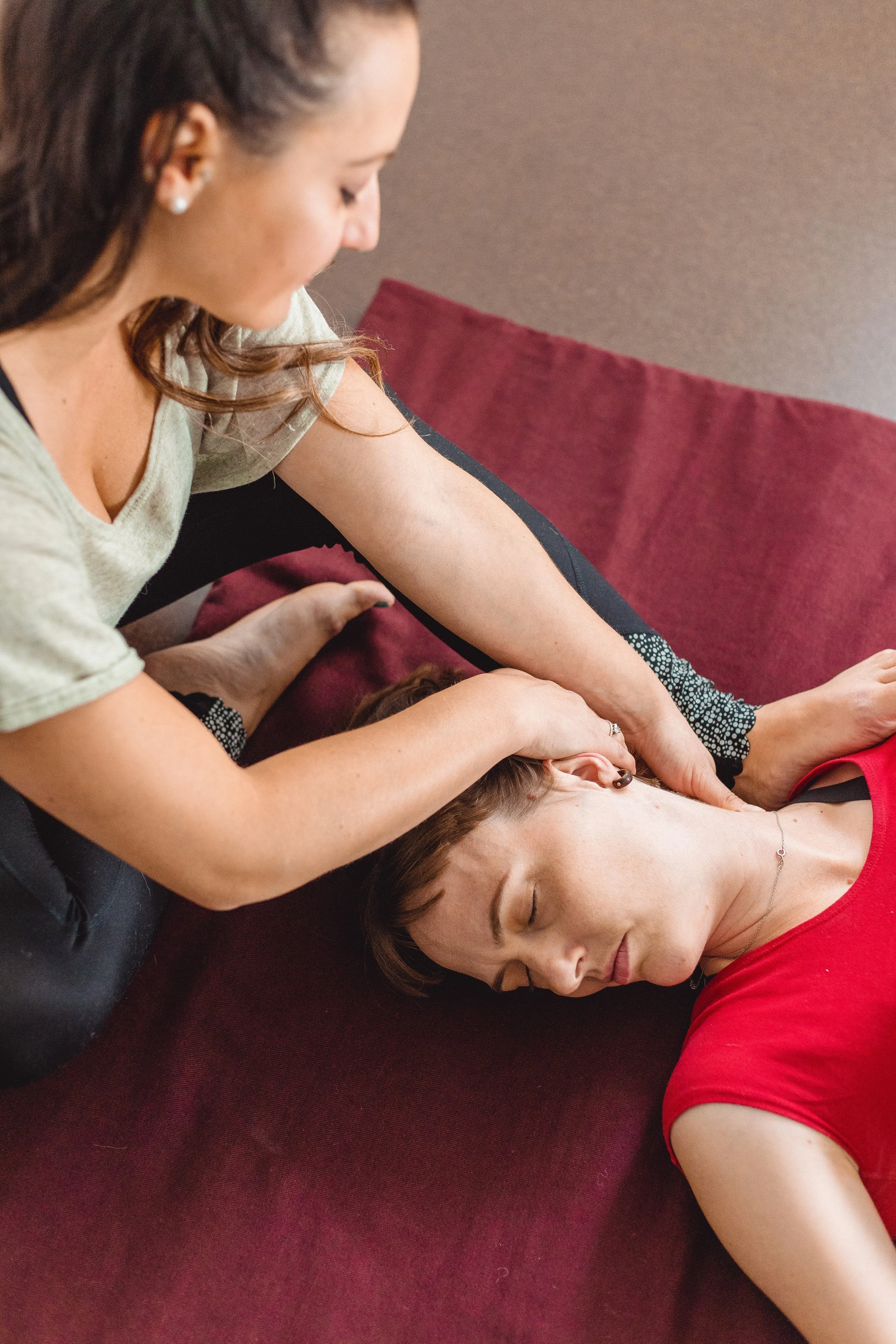 A woman in a black and white dress lying on a red mat, receiving a massage or therapy on her neck and head from another woman with long, wavy brown hair, wearing pearl earrings and a beige top.