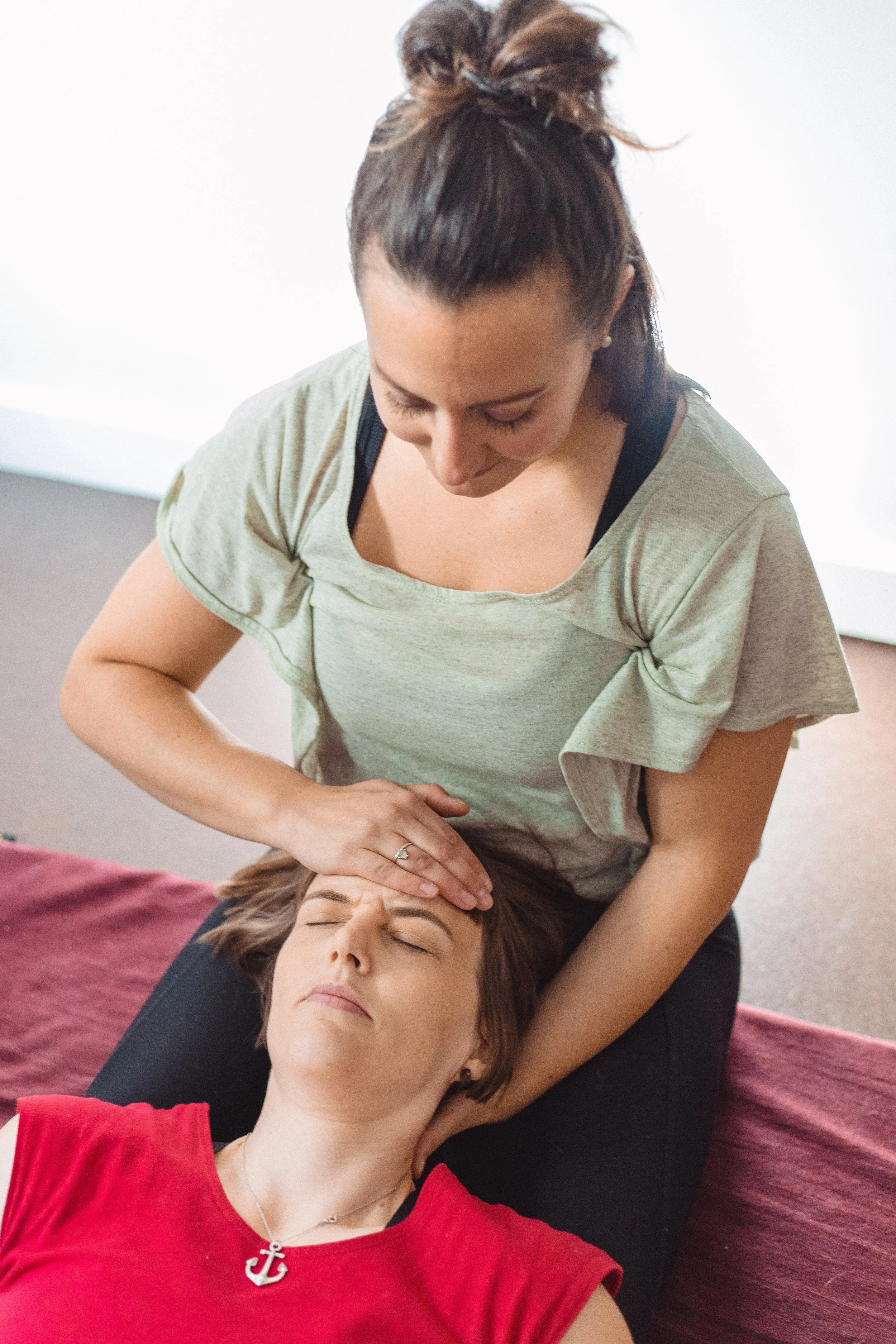 A woman giving a facial massage or therapy to another woman lying on a massage table. The therapist is focused, and the client has her eyes closed.