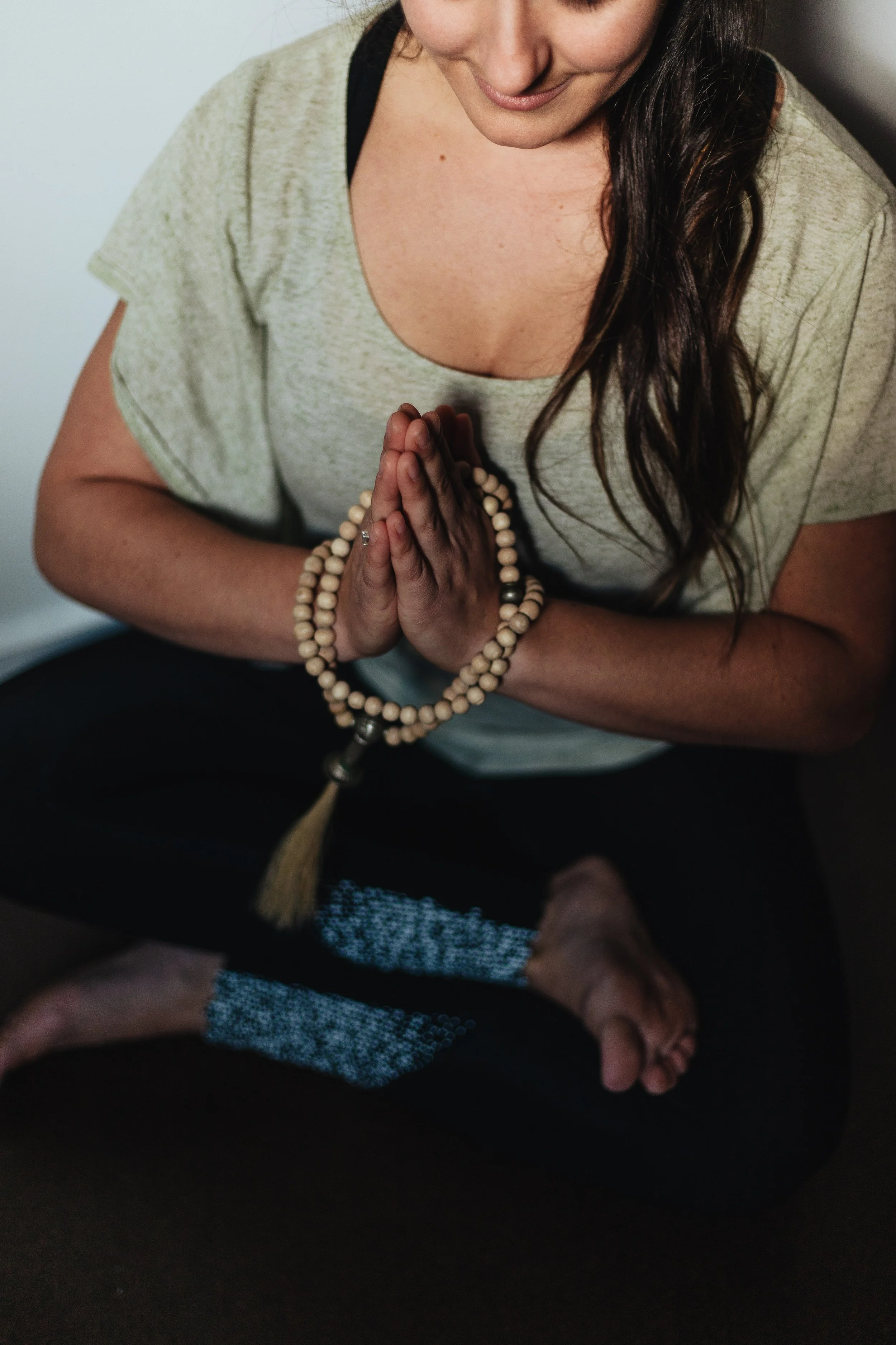 A woman in a beige t-shirt is sitting cross-legged on a dark surface, with her hands pressed together in a prayer position, wearing a beaded bracelet, and smiling down with long hair.