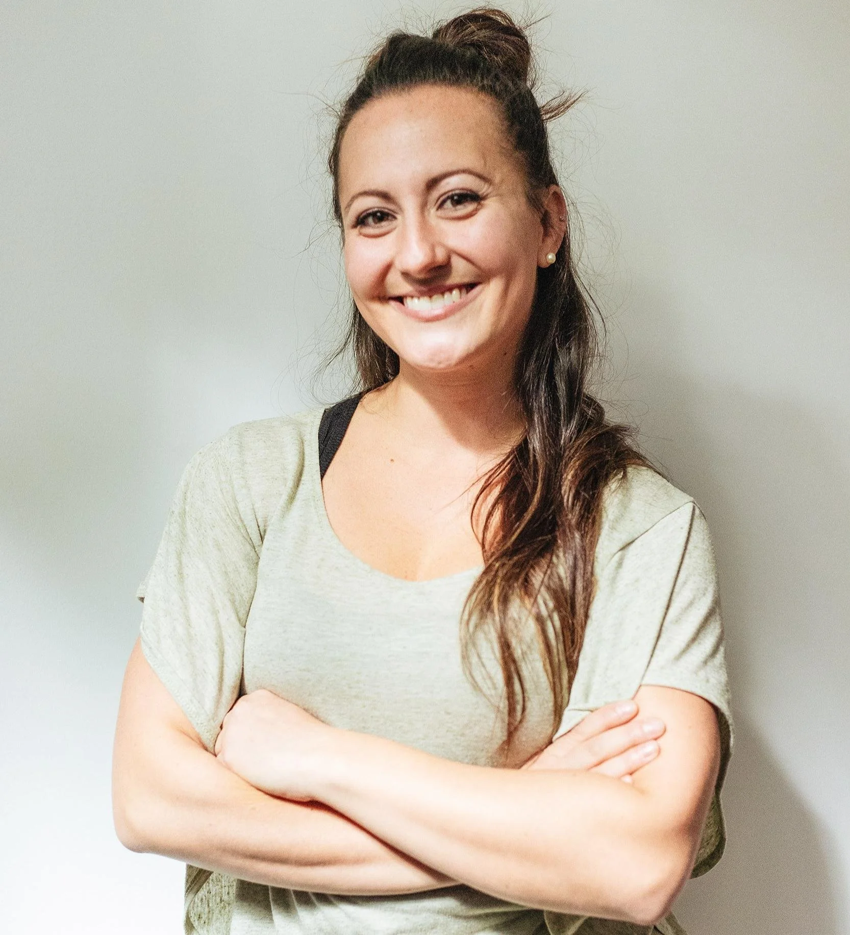 A woman with long brown hair styled in a half-up, half-down hairdo, wearing pearl earrings, a beige top with a black strap visible underneath, smiling with arms crossed against a plain light-colored wall.