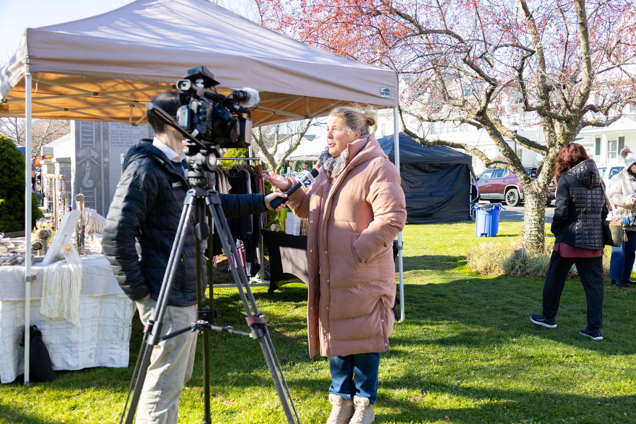 Woman speaking into a news microphone beside a camera operator at an outdoor market