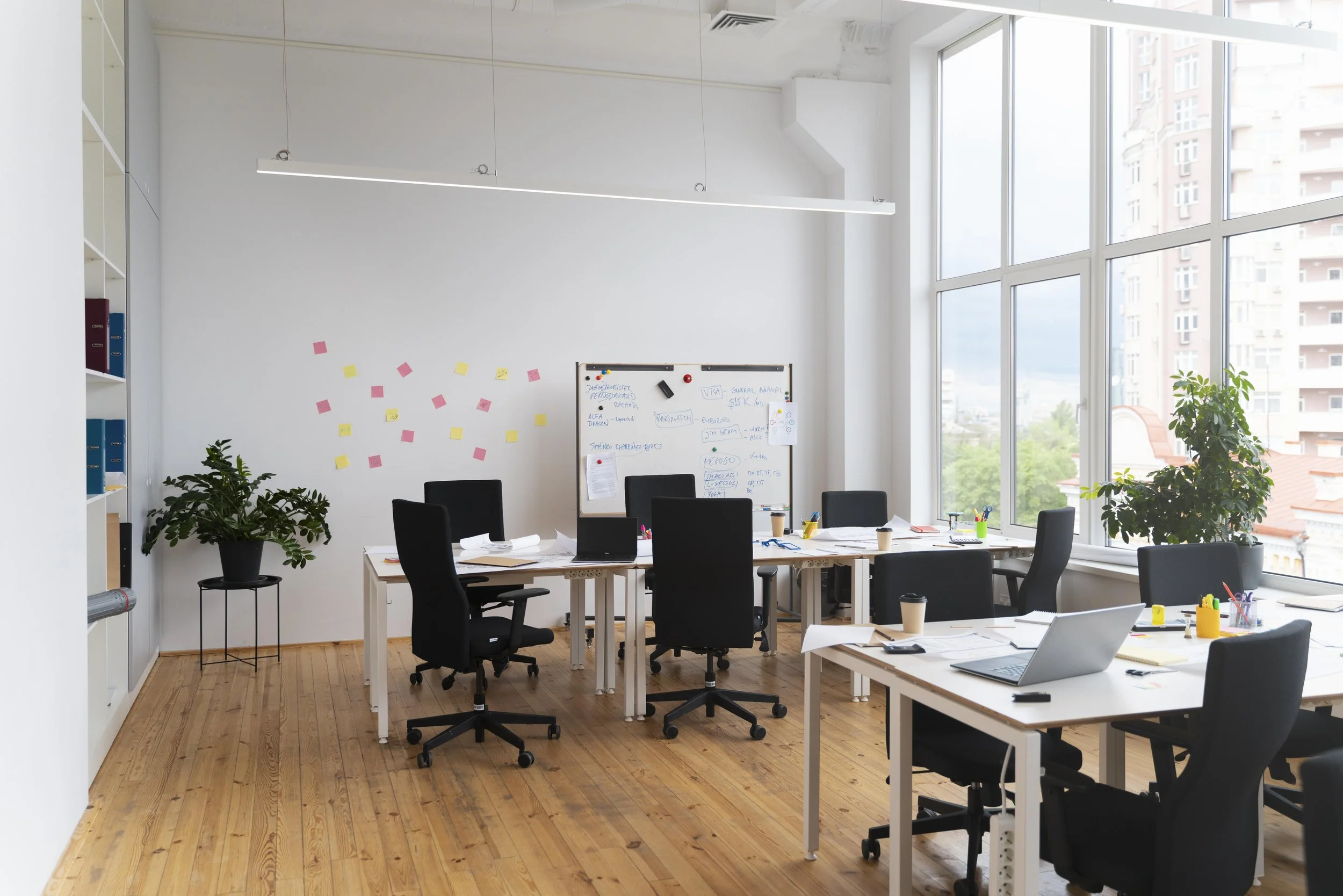 A modern office with large windows letting in natural light, several tables with laptops, papers, and office supplies, black office chairs, potted plants, a whiteboard with notes, and colorful sticky notes on the wall.