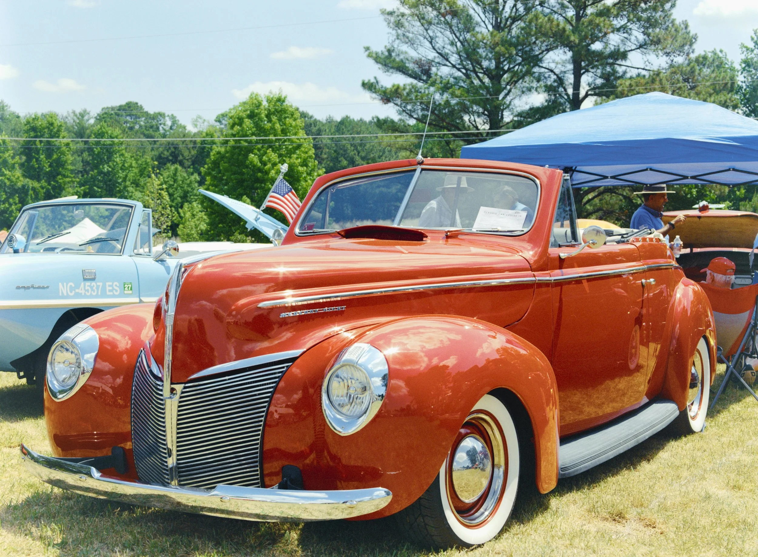 A vintage red car with whitewall tires parked outdoors during a sunny day at a classic car event. In the background, there is a blue canopy tent, people, and lush green trees.