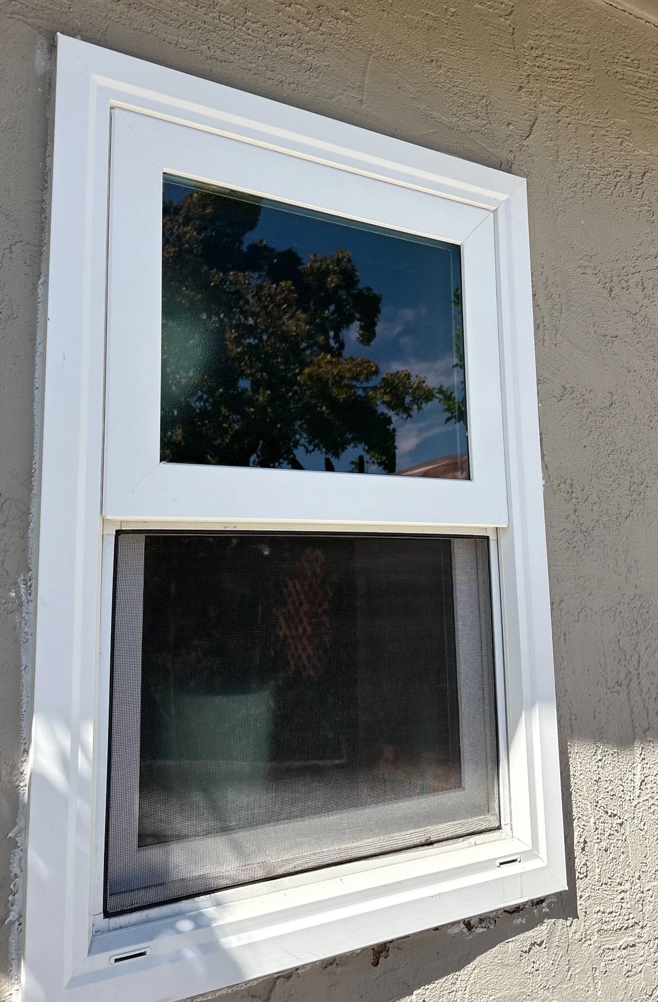 A white-framed window with a screen, set into a beige stucco exterior wall, reflecting clouds and trees in the glass.