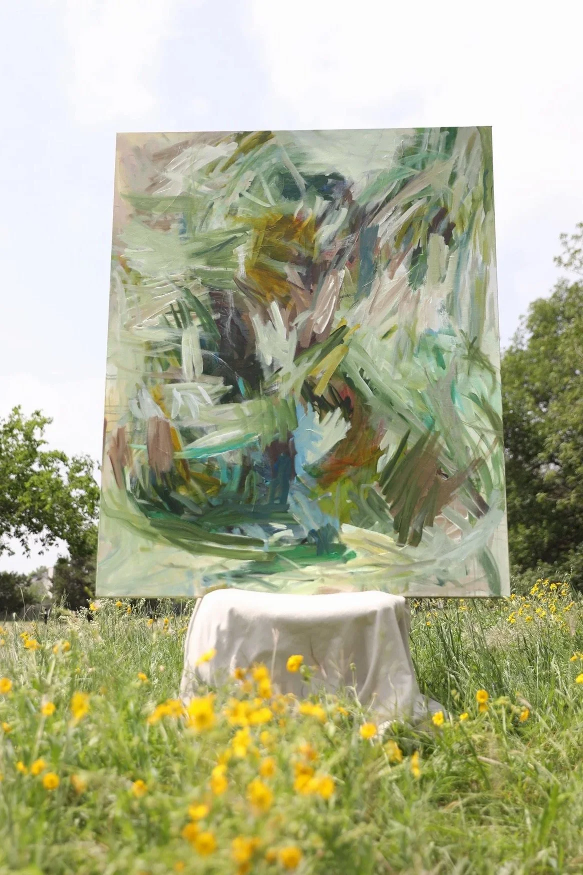 An abstract painting on a large canvas outdoors, with trees and a partly cloudy sky in the background, displayed on a small table in a grassy field with yellow flowers.