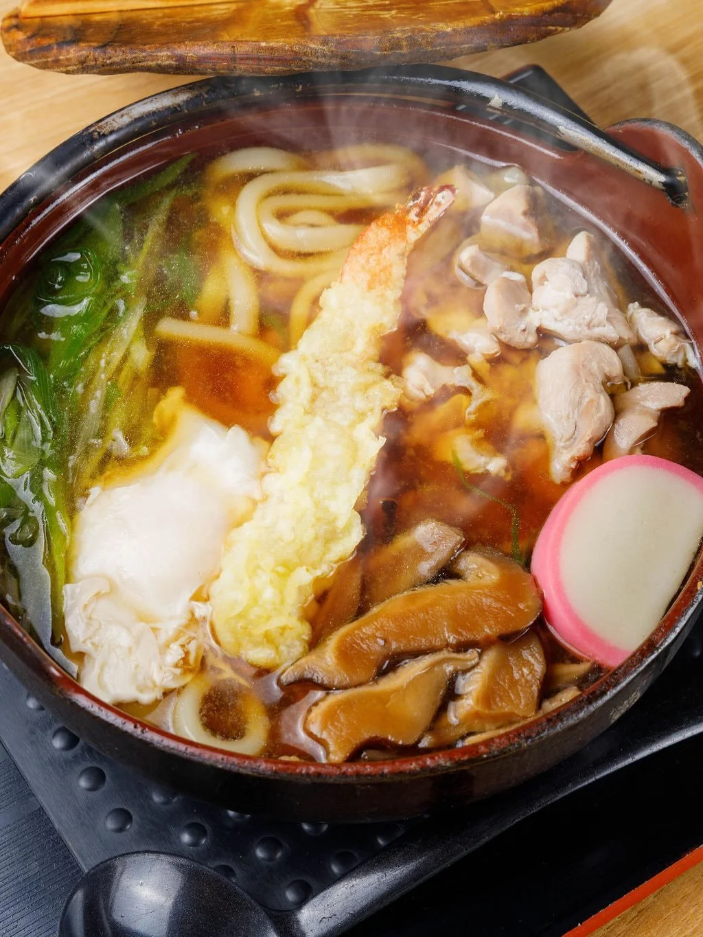 Close-up of a bowl of hot Japanese udon noodle soup with tempura shrimp, sliced fish cake, chicken, mushrooms, spinach, and other ingredients, with steam rising from the soup.