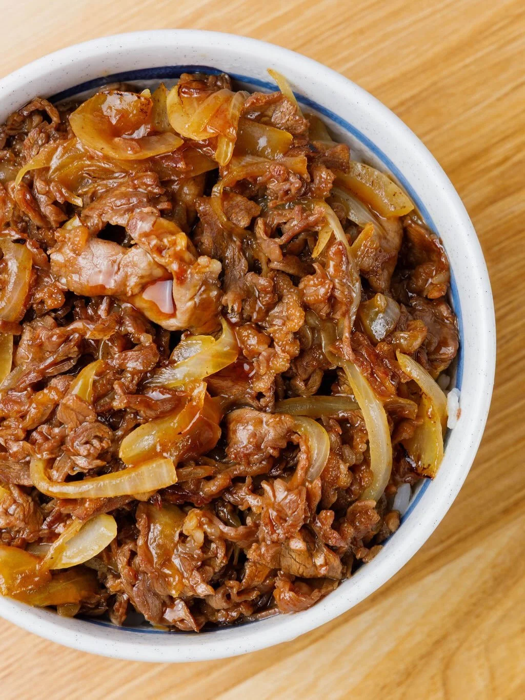 Close-up of a bowl filled with cooked beef, onions, and sauce on a wooden surface.