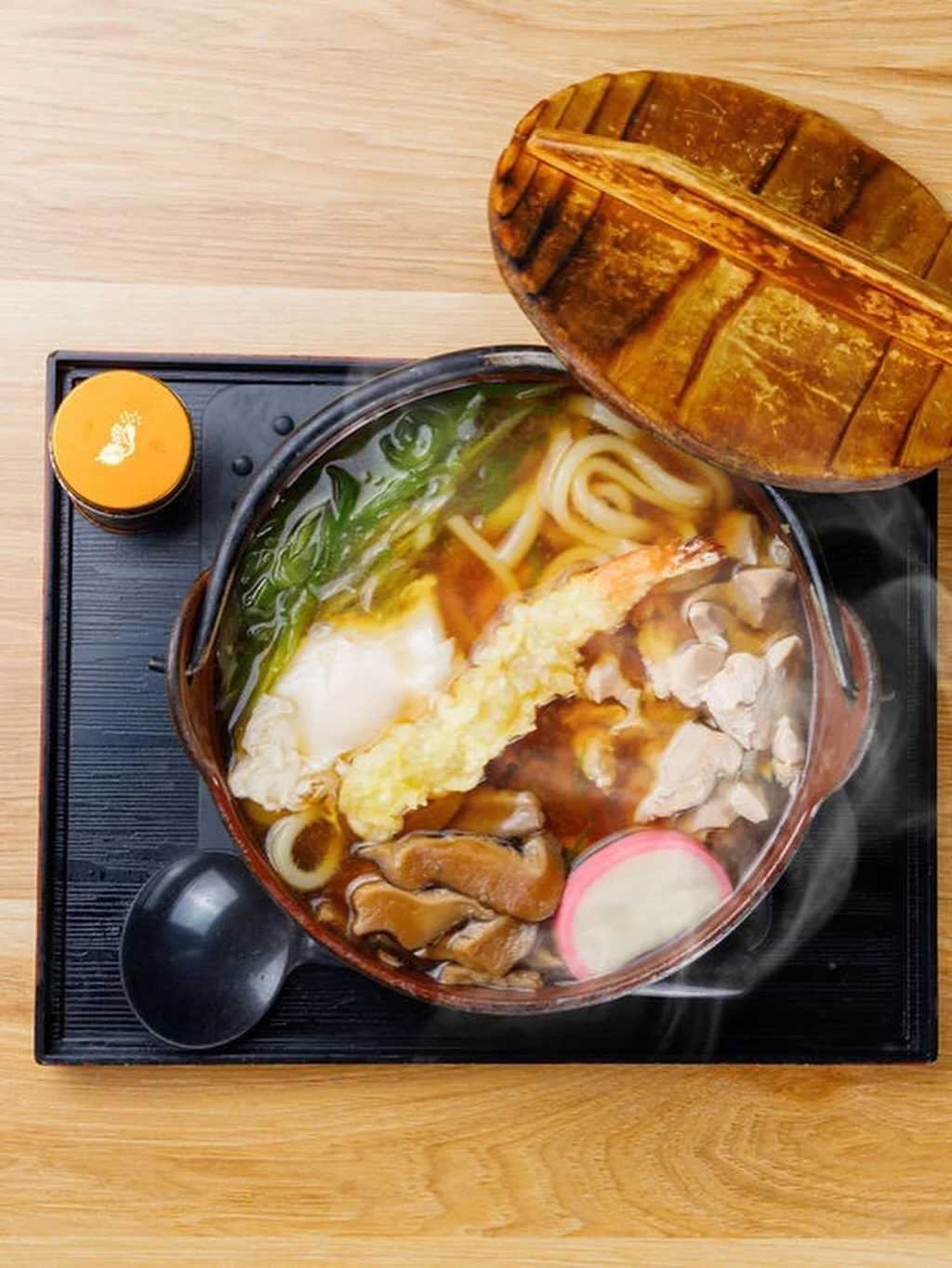 Bowl of ramen with noodles, green onions, mushrooms, tempura, and sliced fish cake on a black tray with a wooden lid, soup spoon, and small container of seasoning.