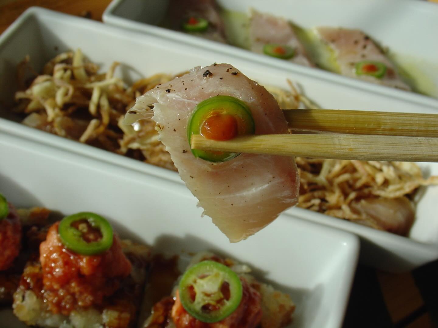Close-up of a piece of raw fish held by chopsticks, garnished with a slice of green chili topped with red sauce, with various Asian dishes in the background.