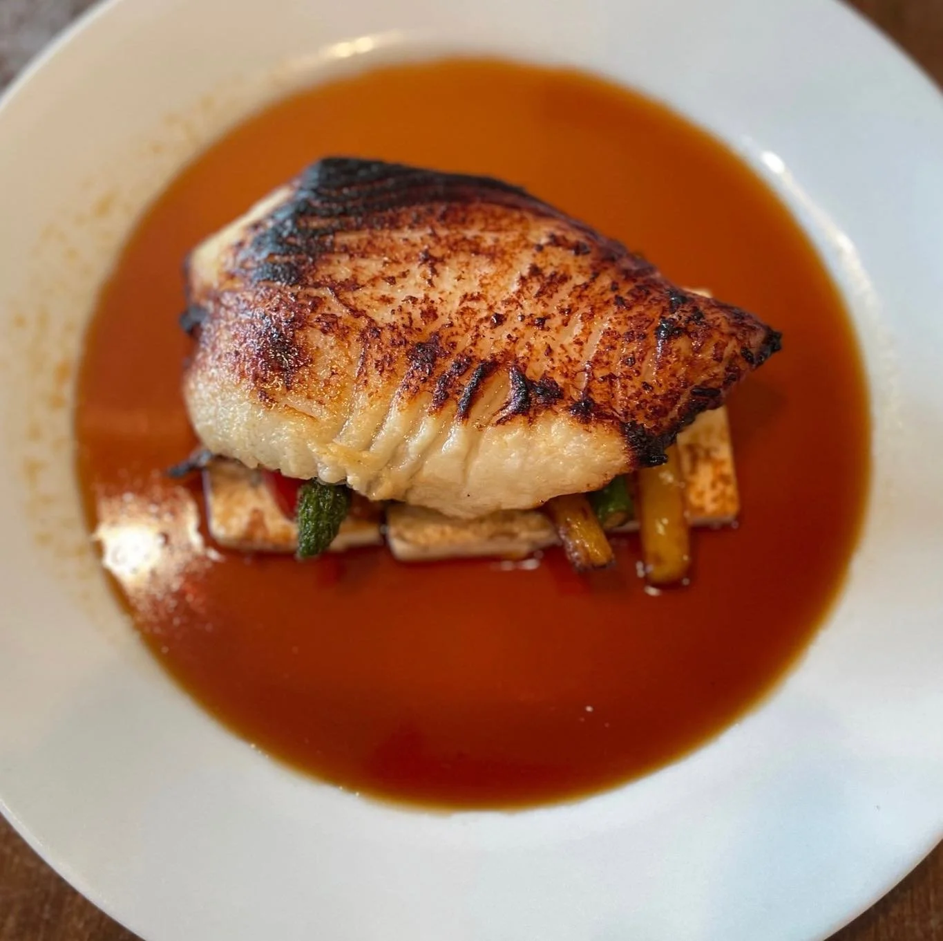 A piece of cooked fish with grill marks served on a bed of vegetables in a brown sauce.