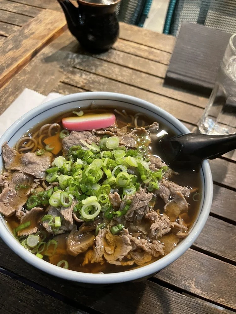 Bowl of ramen topped with sliced beef, chopped green onions, and a slice of fish cake with a pink swirl, on a wooden table with a glass of water and chopsticks.