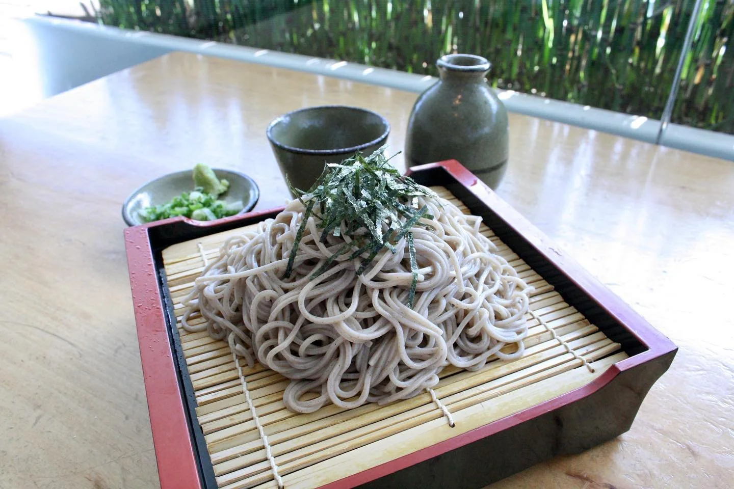 A serving of soba noodles topped with shredded nori seaweed, served on a bamboo mat with side condiments of wasabi and green onions, accompanied by a small cup and a ceramic sake bottle on a wooden table.