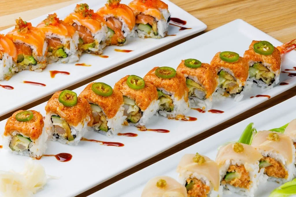 Three plates of sushi rolls garnished with sauce and sliced jalapenos, on a wooden surface.