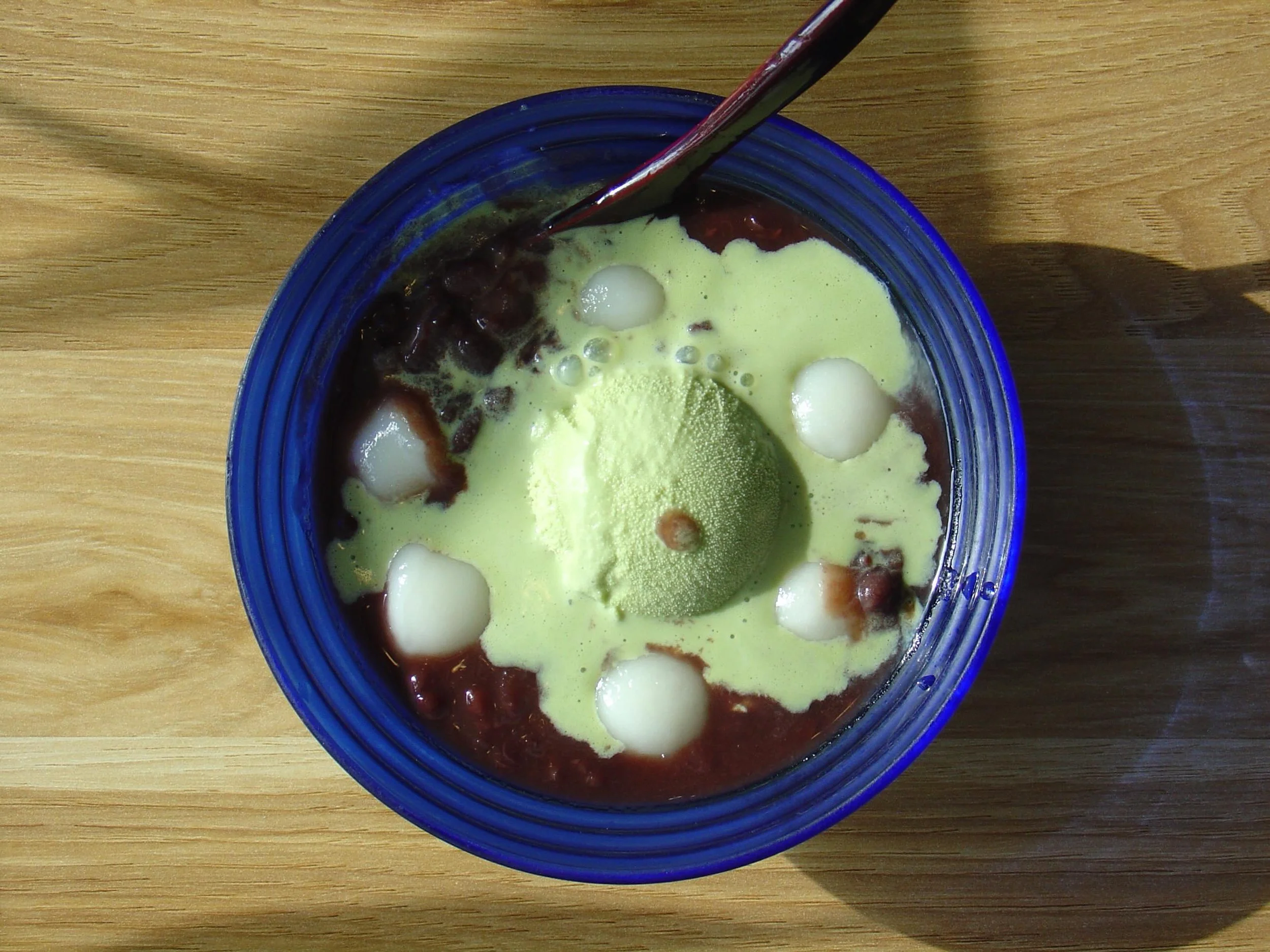 A bowl with green tea and small white balls, topped with red beans and a dollop of cream, on a wooden surface.