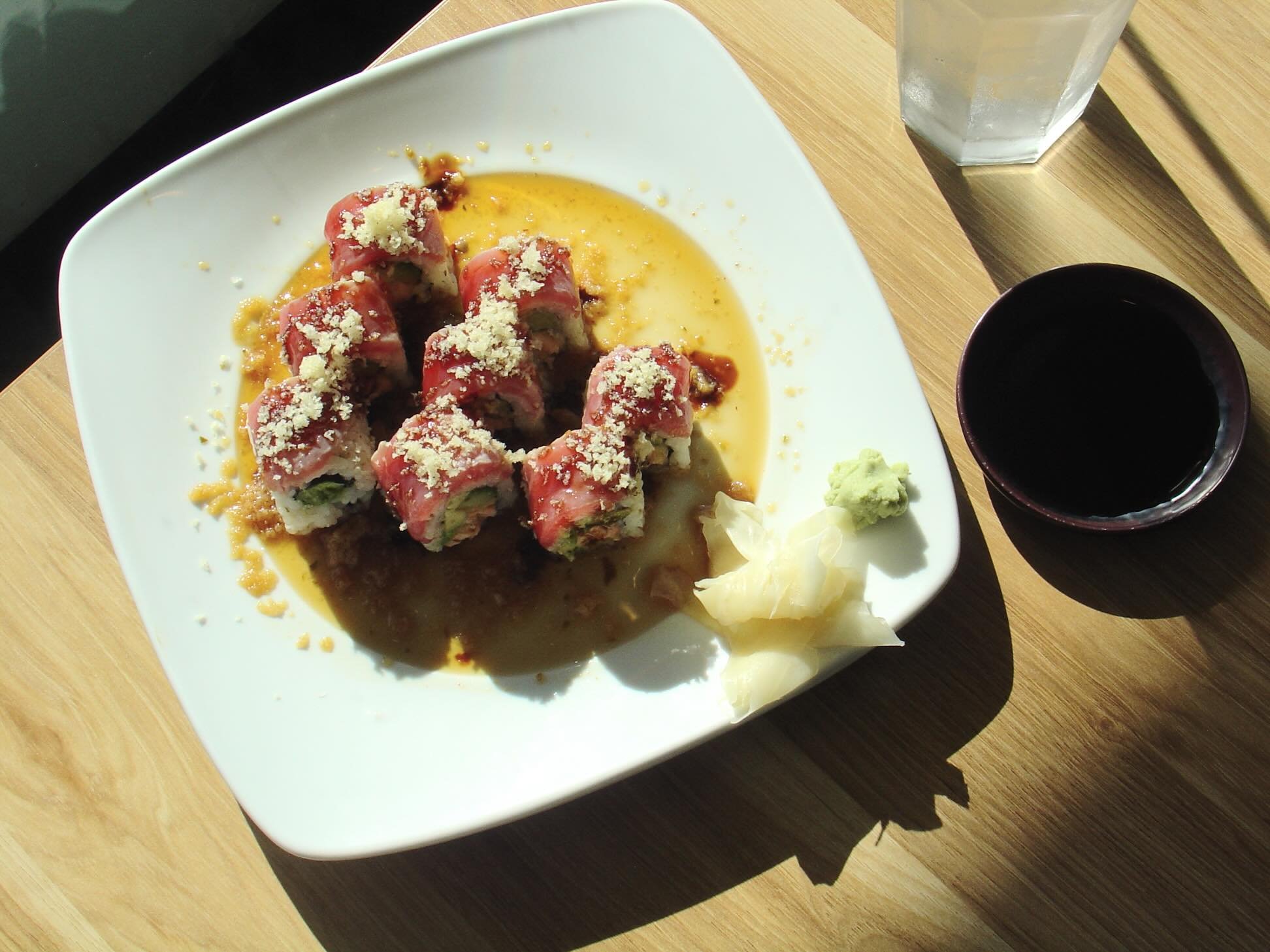 A white plate of sushi rolls topped with sliced meat, drizzled with sauce, and sprinkled with cheese, accompanied by wasabi and pickled ginger. A small black bowl of soy sauce, a glass of water, and a cup of dark sauce or tea are on the wooden table.