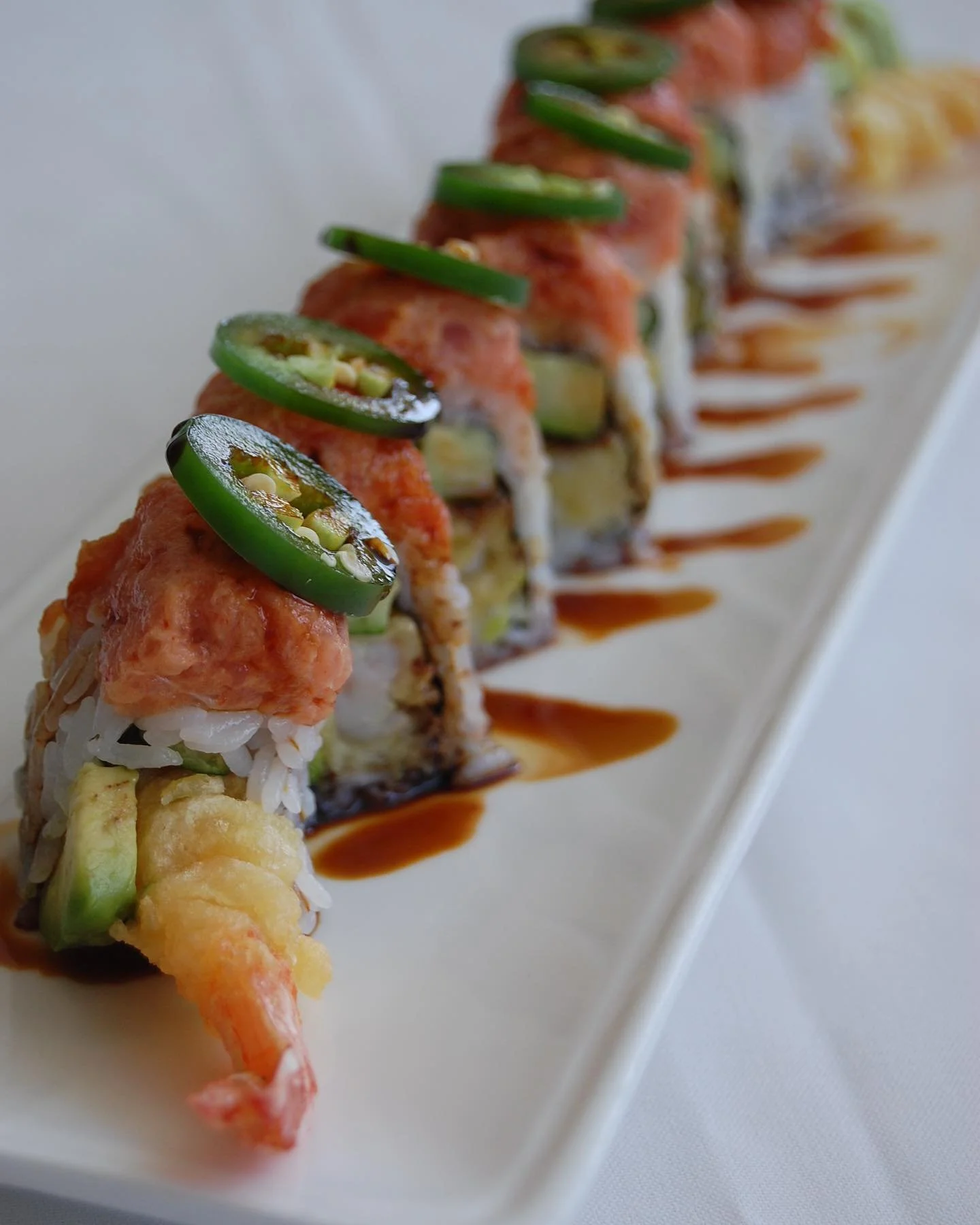 Close-up of a sushi roll topped with sliced jalapeño peppers, drizzled with sauce, served on a white rectangular plate.