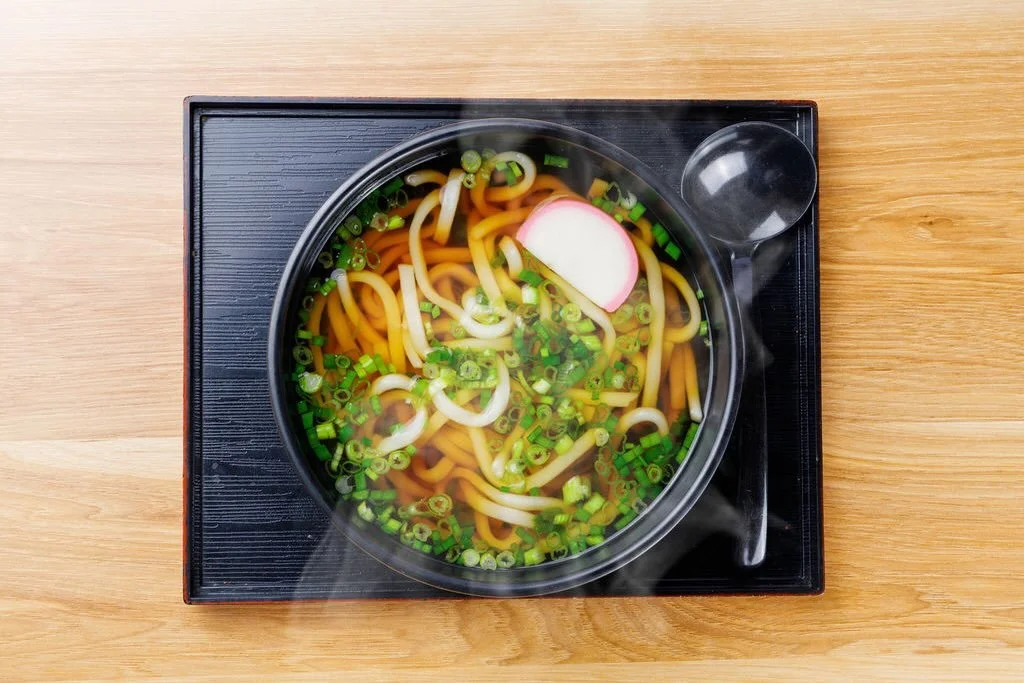 Bowl of steaming udon noodle soup with sliced green onions and fish cake on black tray