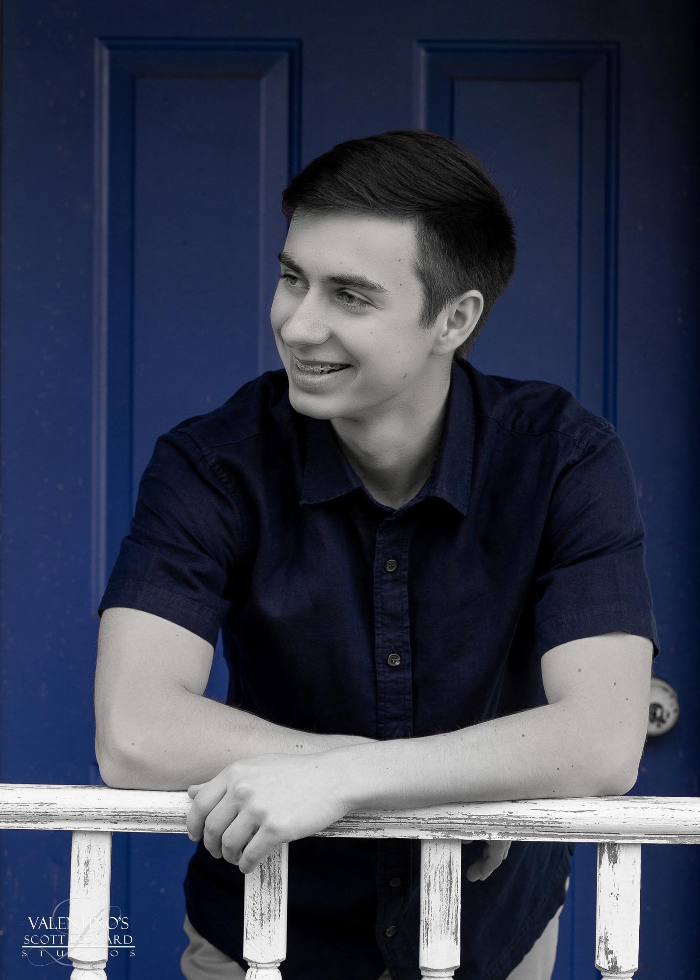 Young man with dark hair and wearing a dark shirt, leaning on a white railing in front of a blue door.
