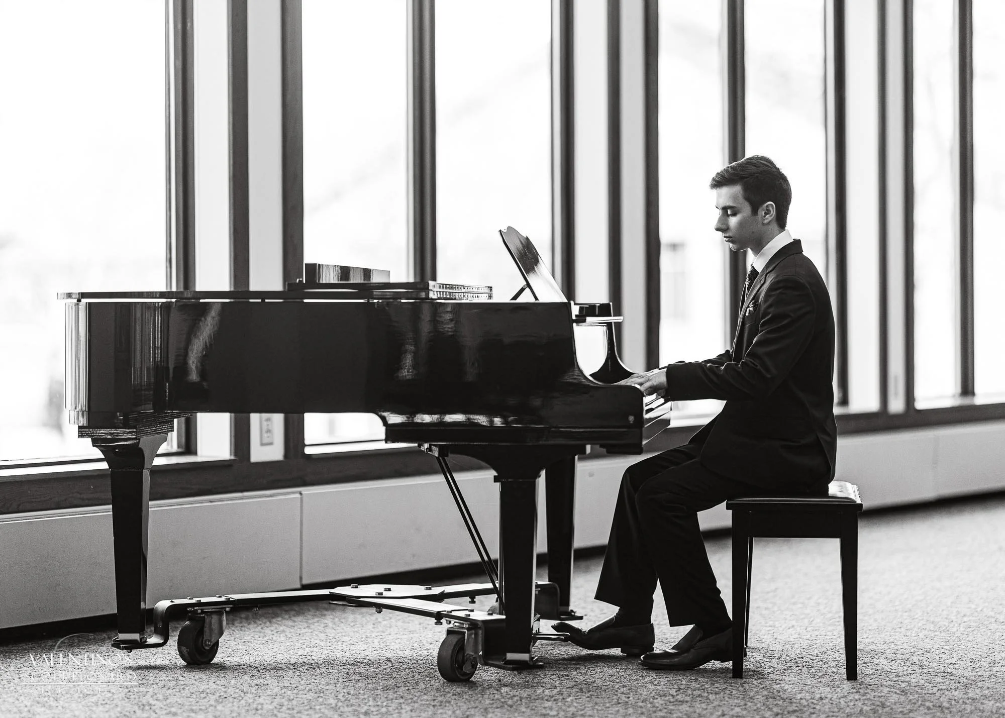 A young man in a suit playing a grand piano indoors, sitting on a bench with a music stand nearby, with large windows in the background.