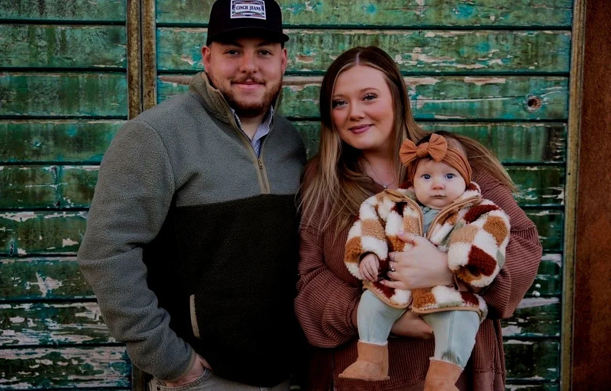 A family group of three: a smiling man with a beard and a black cap, a woman holding a baby girl, standing in front of a weathered green wooden background.