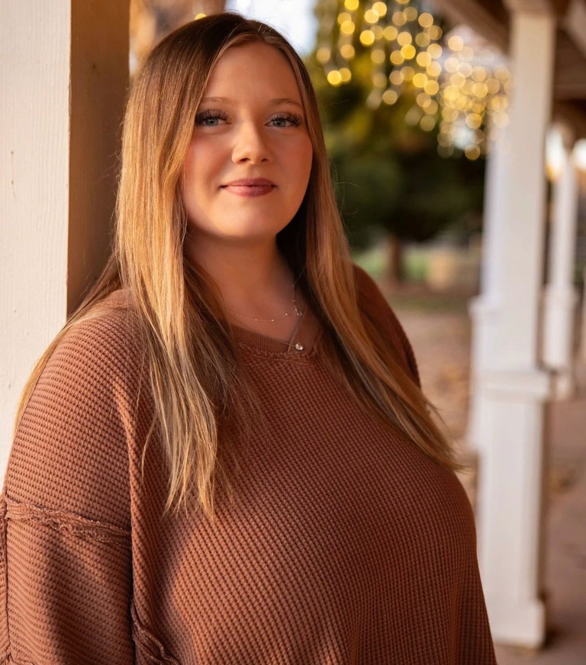 A woman with long, light brown hair and blue eyes standing outdoors near a porch, smiling at the camera with blurred holiday lights and trees in the background, wearing a brown sweater and a necklace.
