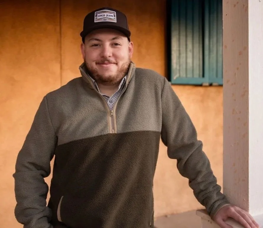 A young man with a beard, wearing a cap and a two-tone fleece jacket, stands indoors against a warm-colored wall with a window behind him, smiling at the camera.