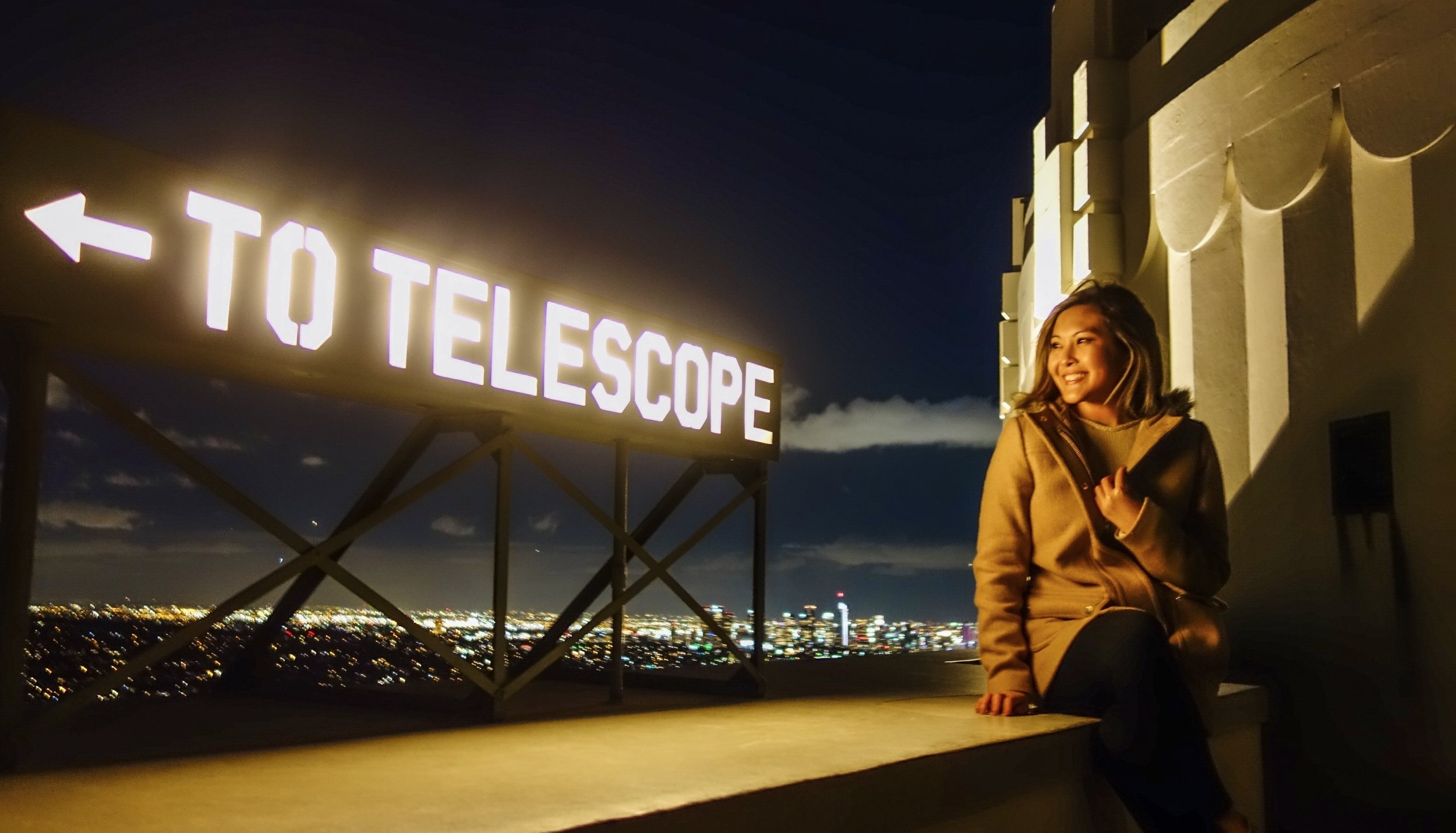 A woman smiling and sitting at night near a lit-up sign that reads 'TO TELESCOPE' with a cityscape in the background.