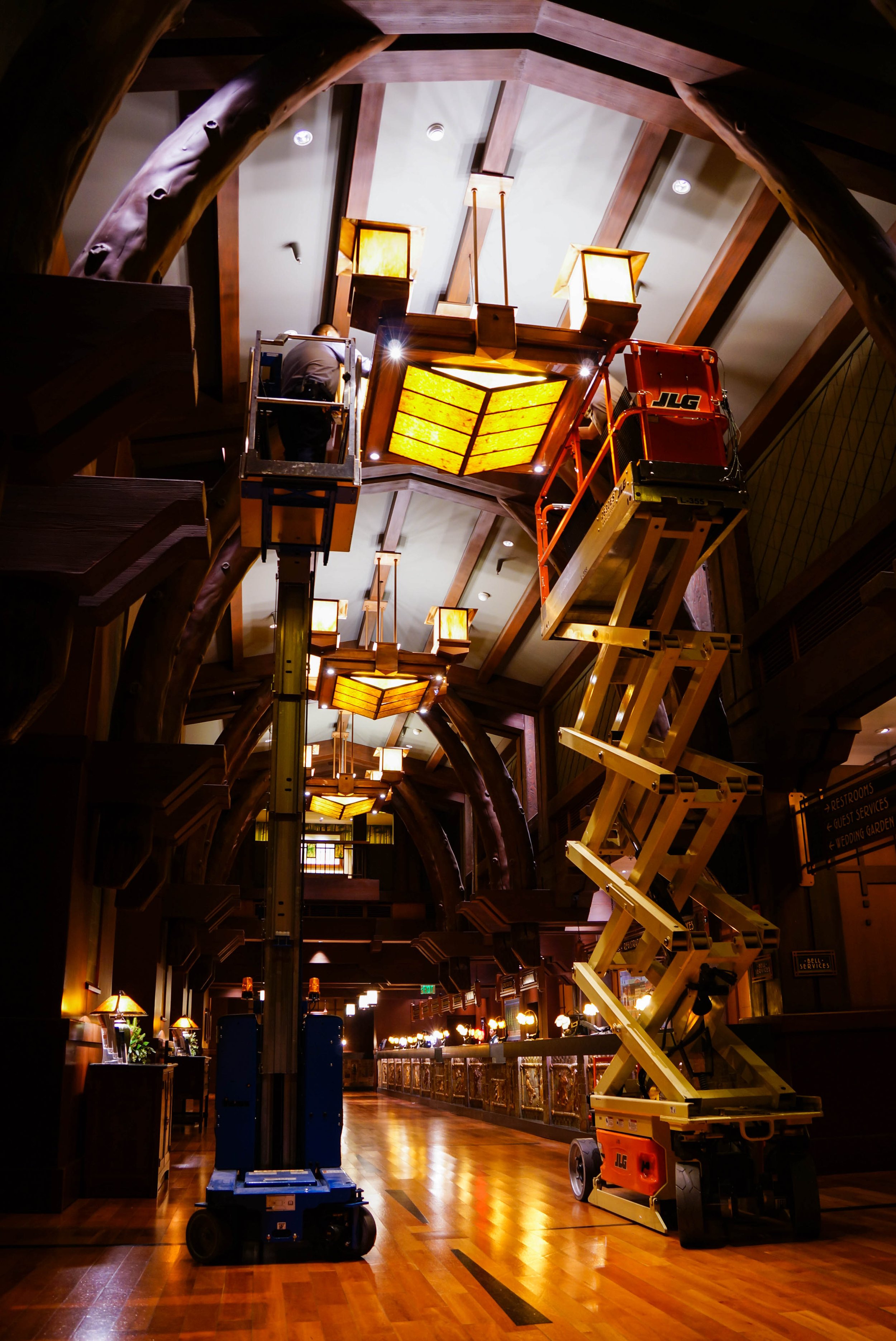 Interior of a building with wooden beams, warm lighting, and a high ceiling. Two scissor lifts are positioned near the center, with one worker on a lift. The space is decorated with lantern-style light fixtures hanging from the ceiling, and the floor
