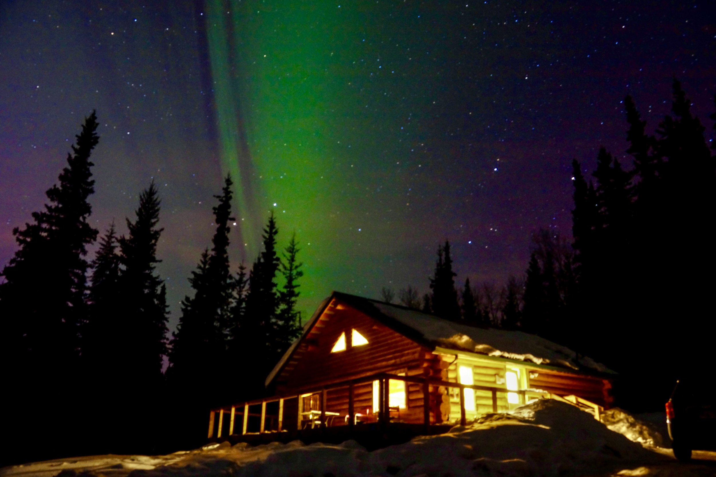 Night sky with northern lights and numerous stars above a cabin surrounded by trees and snow.