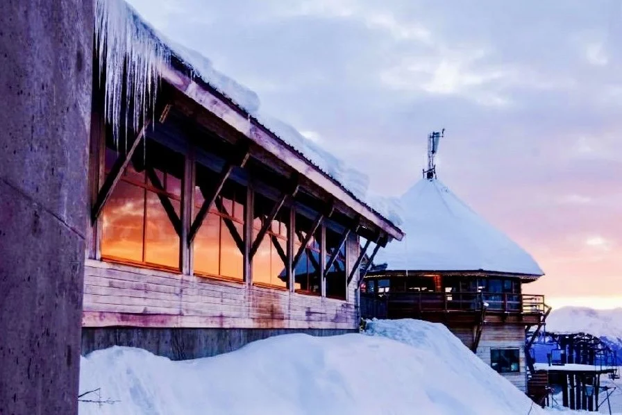 Snow-covered wooden building with icicles hanging from the roof, overlooking a sunset sky.