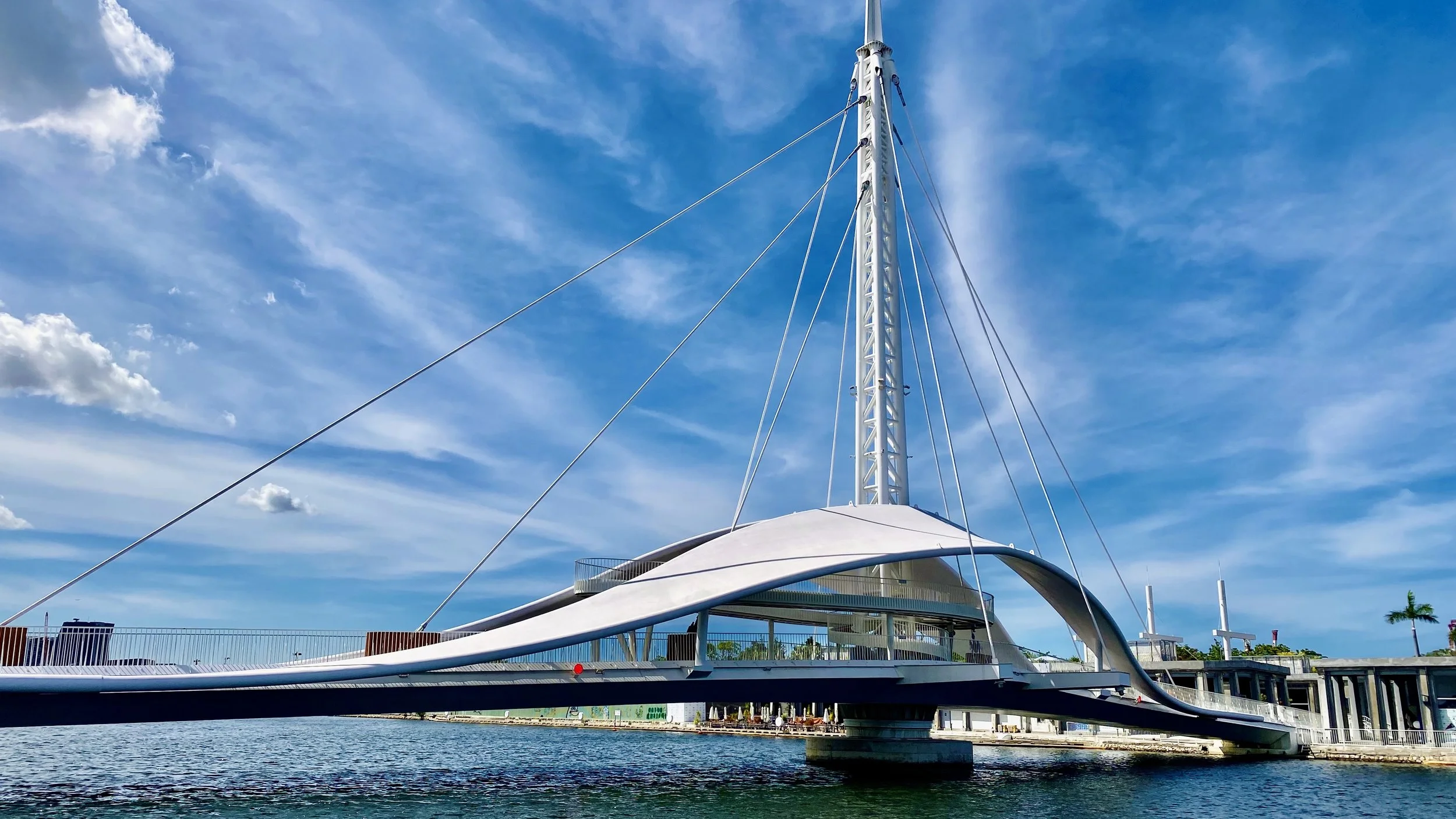 A modern yacht with a white, futuristic design, featuring a tall mast and sleek curves, docked at a marina under a partly cloudy blue sky.
