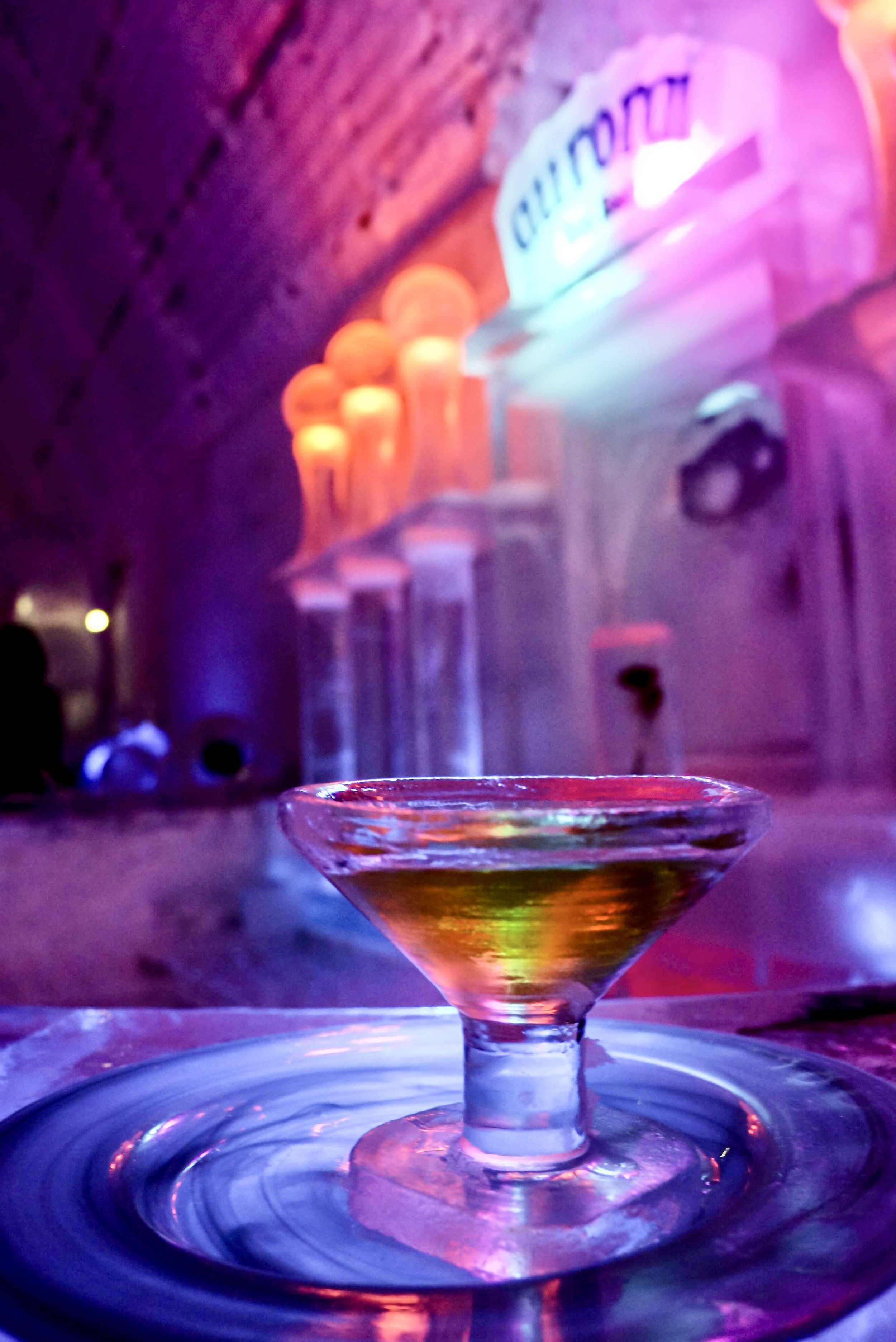 Close-up of a martini glass filled with a colorful drink on a reflective surface, with blurred neon lights and signage in the background, creating a vibrant atmosphere.
