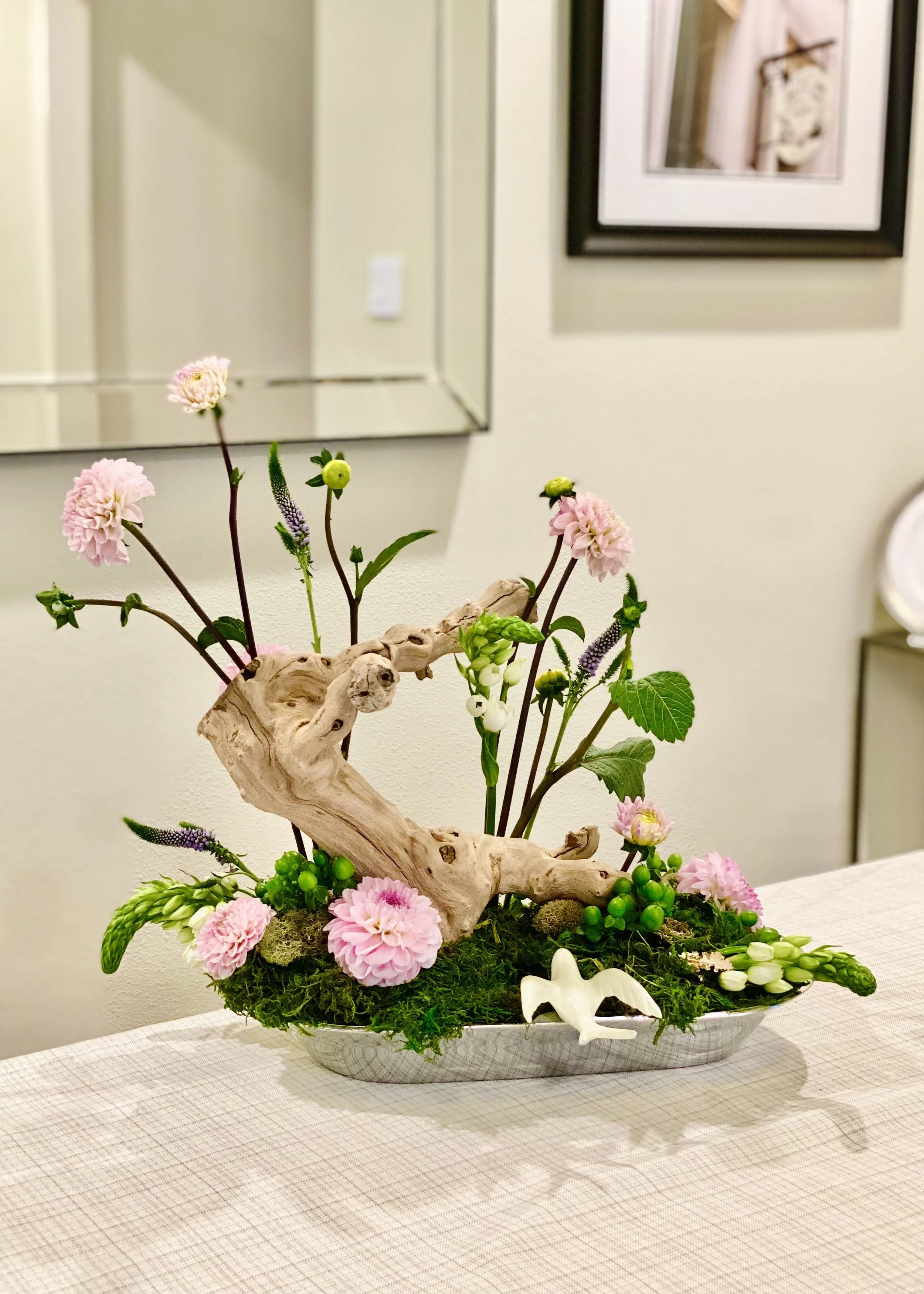 A floral arrangement in a white ceramic bowl with pink flowers, green foliage, and a twisted piece of driftwood, placed on a table with a beige tablecloth and a star-shaped ornament.