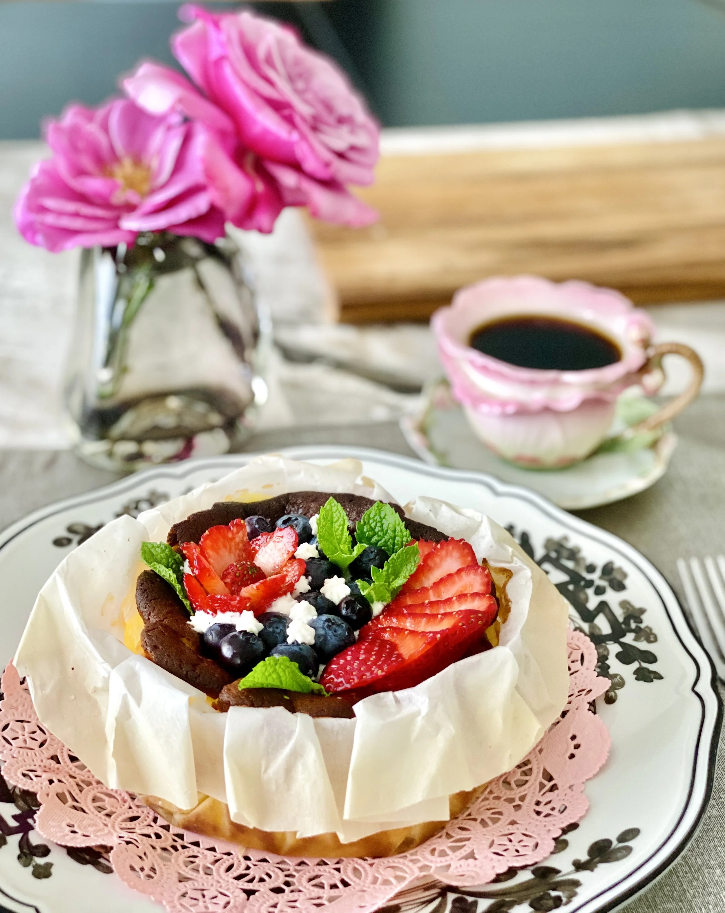 A dessert topped with fresh strawberries, blueberries, and mint leaves on a decorative white and black plate with a pink paper doily.