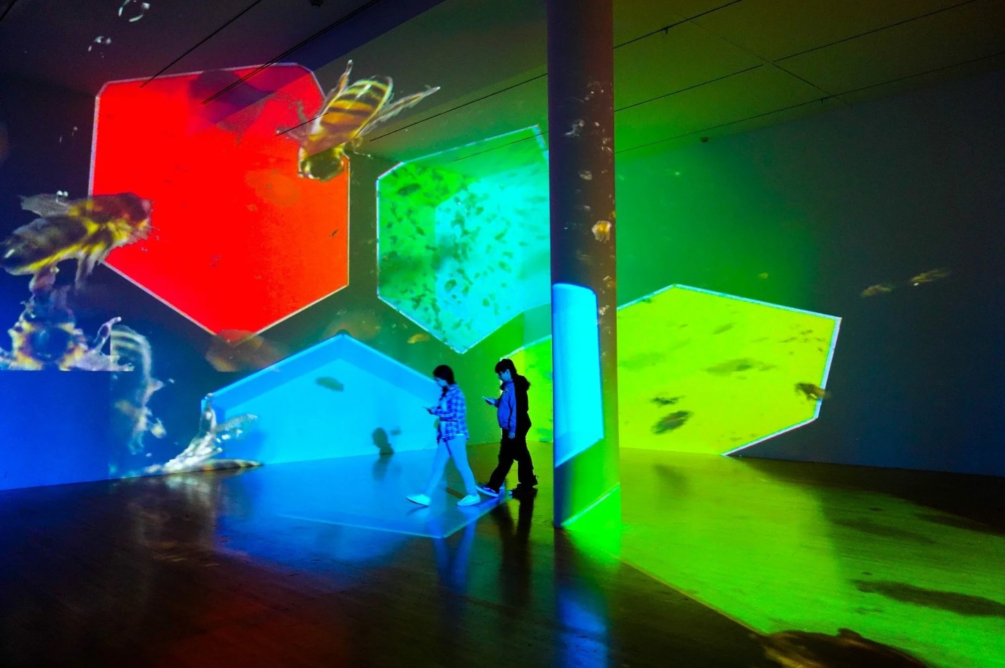 People walking in a dark room with neon-colored geometric shapes projected on the walls and ceiling.