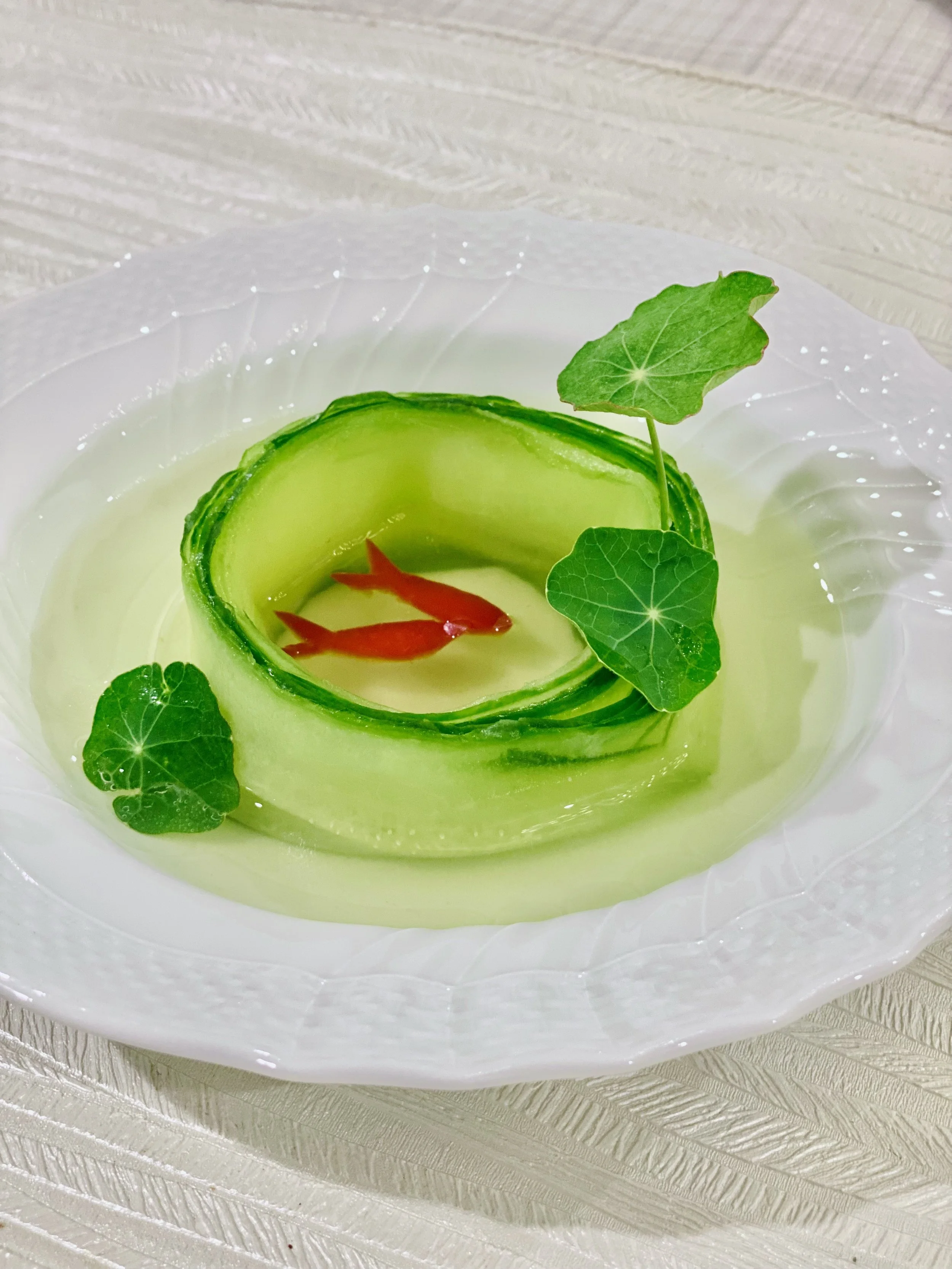 A white plate with a decorative rim, holding a cucumber slice with a small red fish and decorated with green leaves.