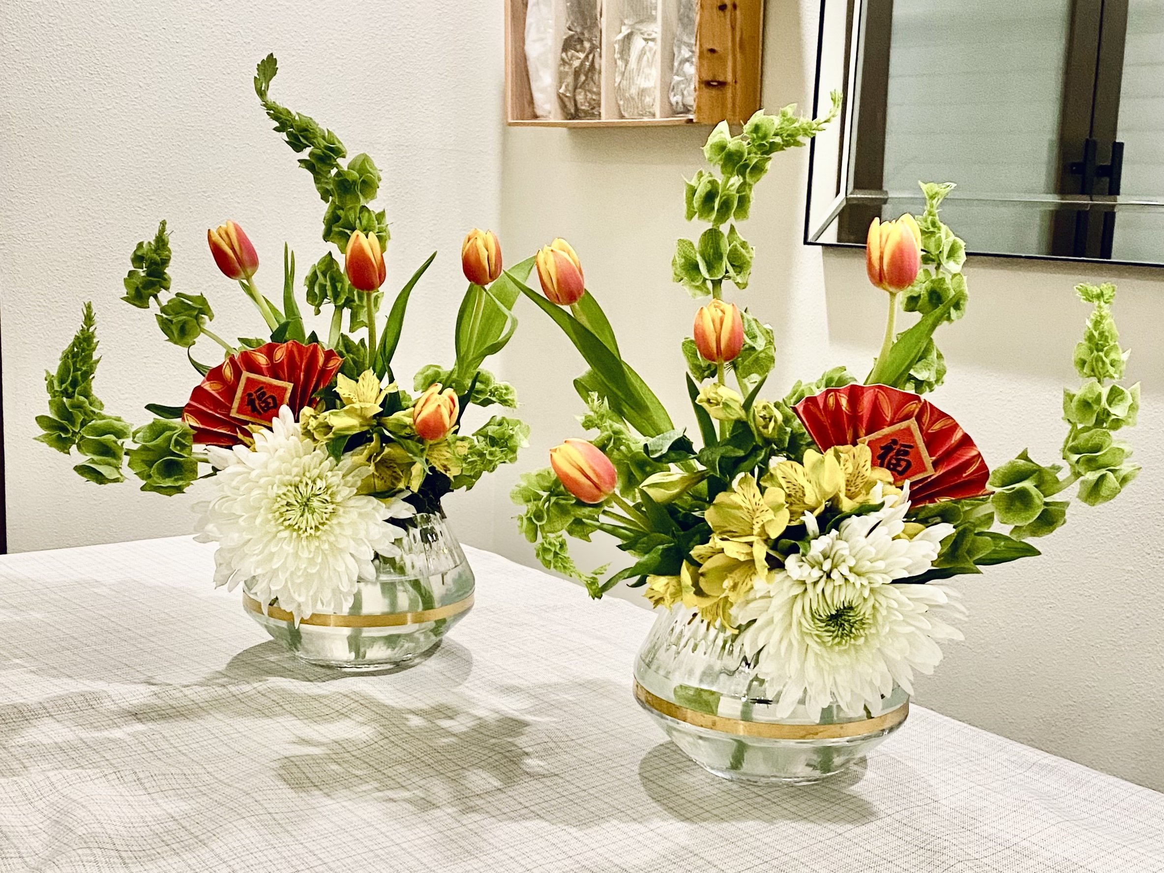Two glass vases with colorful flower arrangements including white, yellow, orange, and red flowers, placed on a white tablecloth in front of a beige wall with a mirror and wooden shelf.