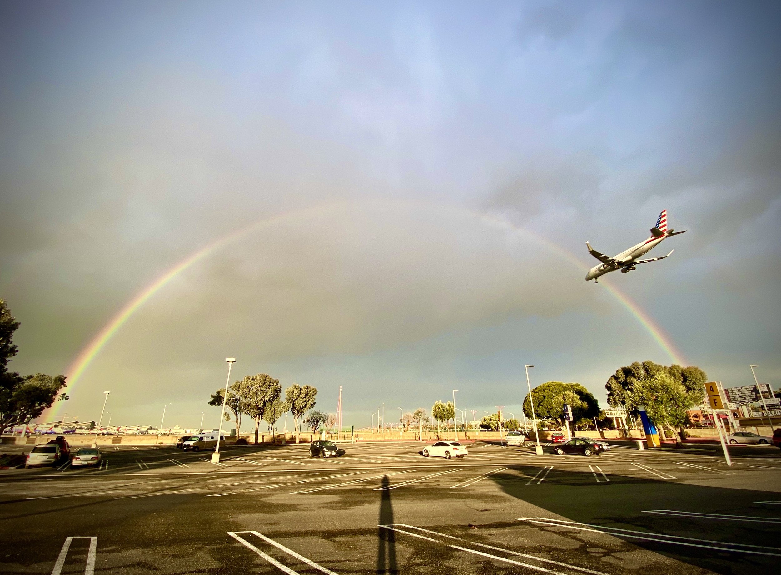 An airplane with an American flag painted on its tail is flying in the sky over a parking lot, with a rainbow visible in the background and trees lining the area.