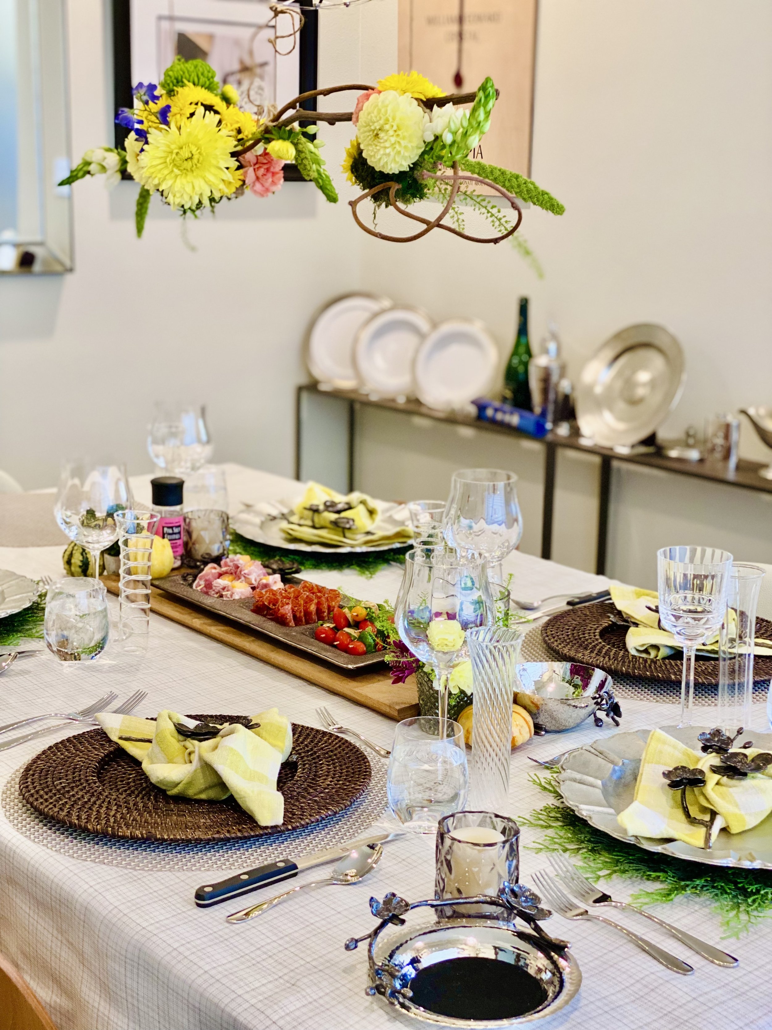 A decorated dining table with plates, glasses, cutlery, and napkins, featuring a centerpiece and a platter of assorted foods, with a hanging floral arrangement above and a sideboard in the background holding additional dishes and decorative items.