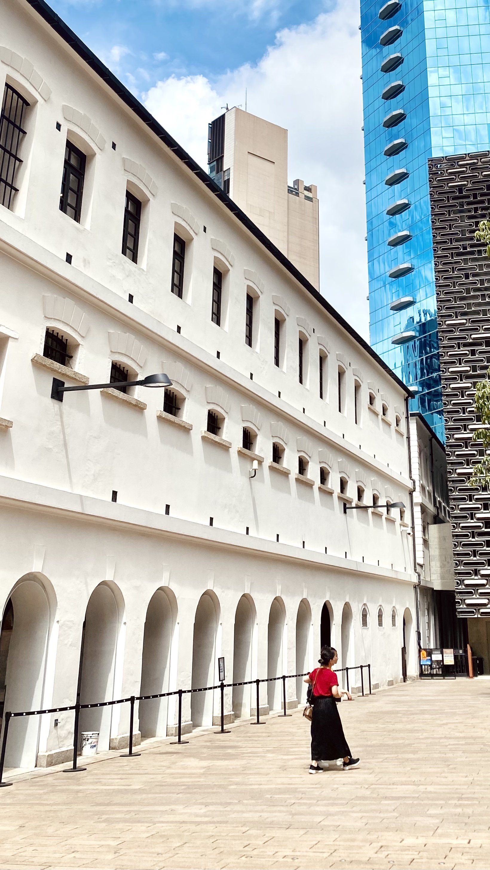 A woman in a red top and black skirt walking past a white building with arched windows and black barred windows in an urban cityscape with modern tall buildings and a blue sky.