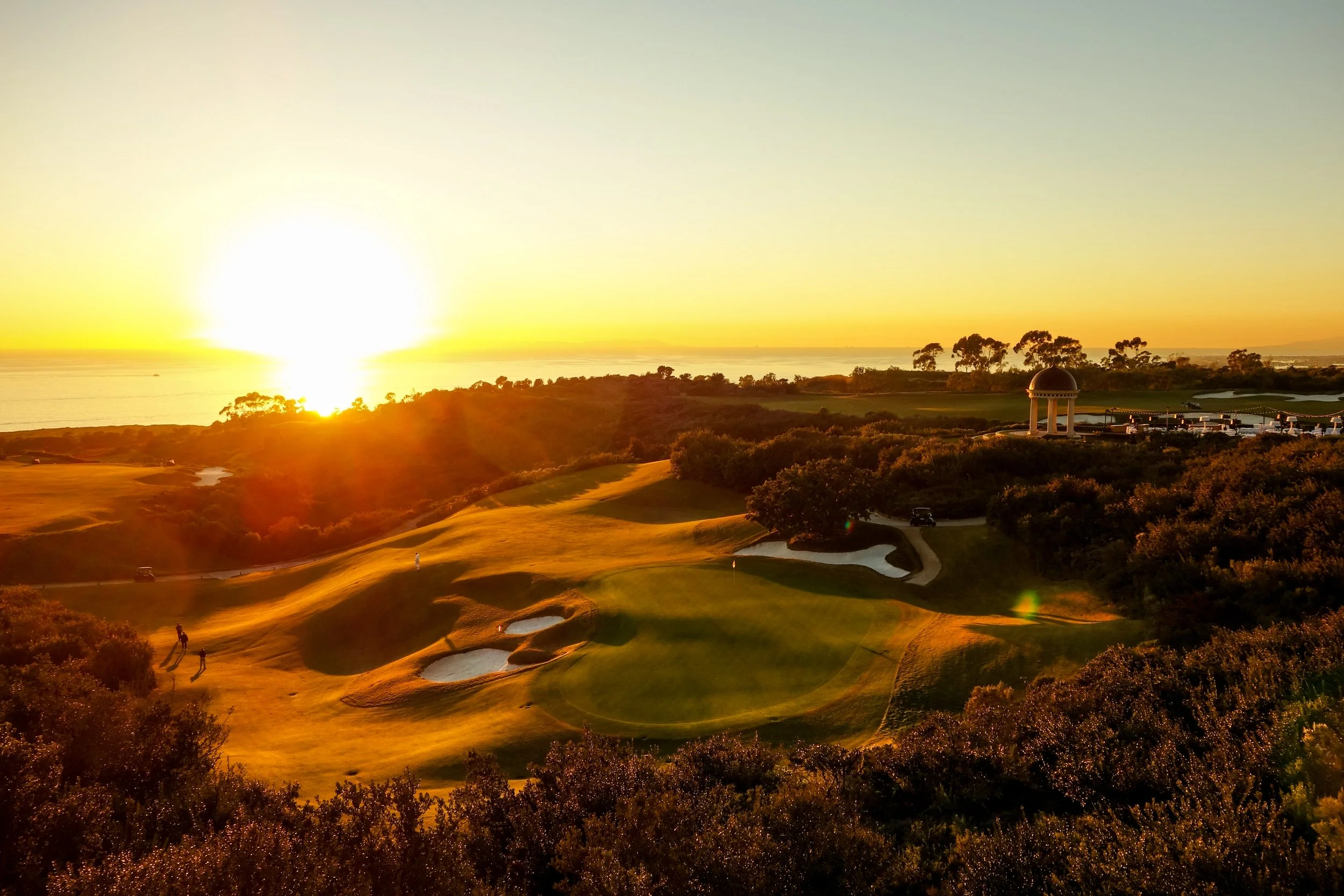 A sunset over a golf course near the ocean, with trees and sand traps visible.