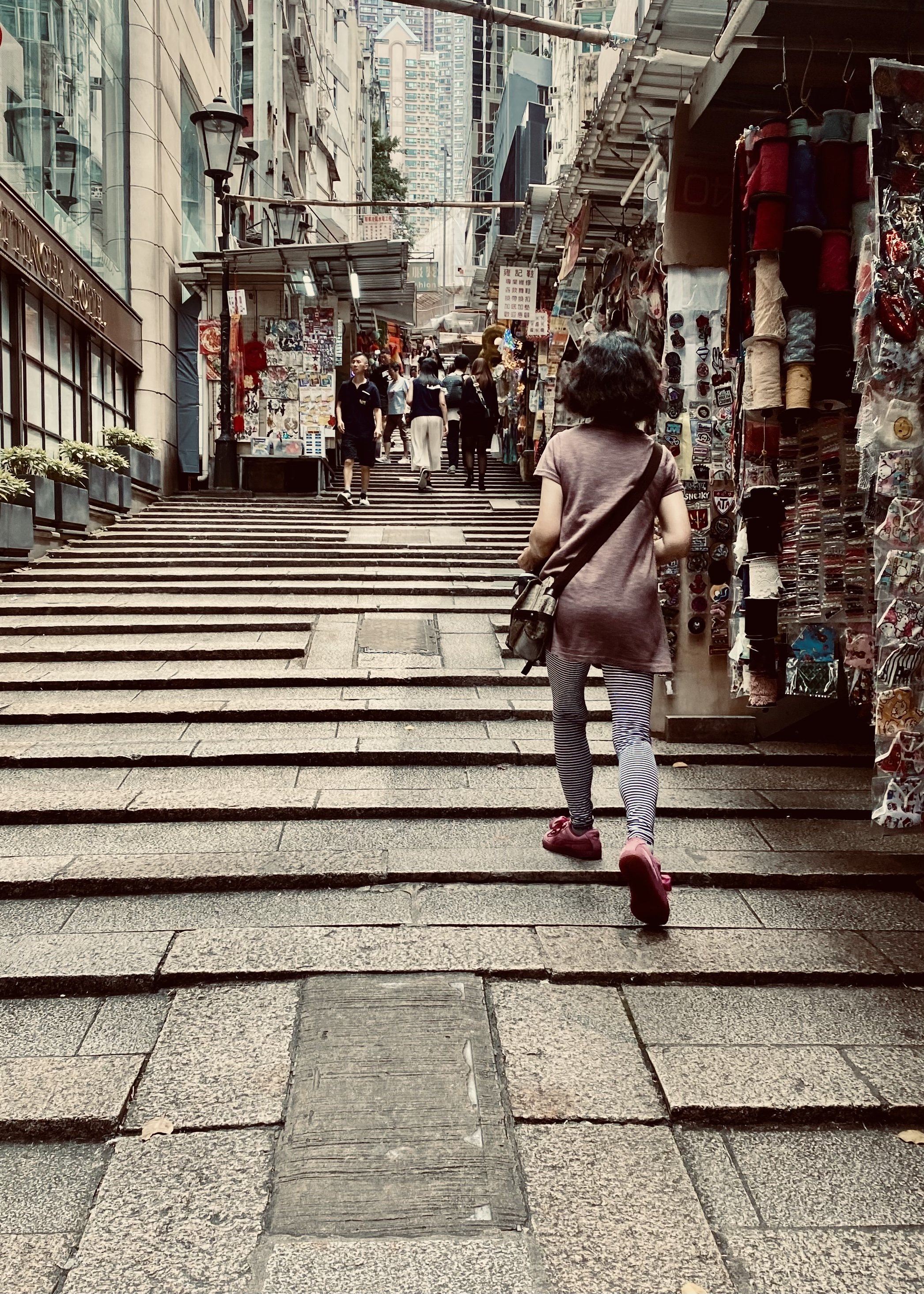 A woman is walking up a set of stone stairs on a city street, with shops and market stalls on either side. There are other people walking up and down the stairs, and high-rise buildings are visible in the background.