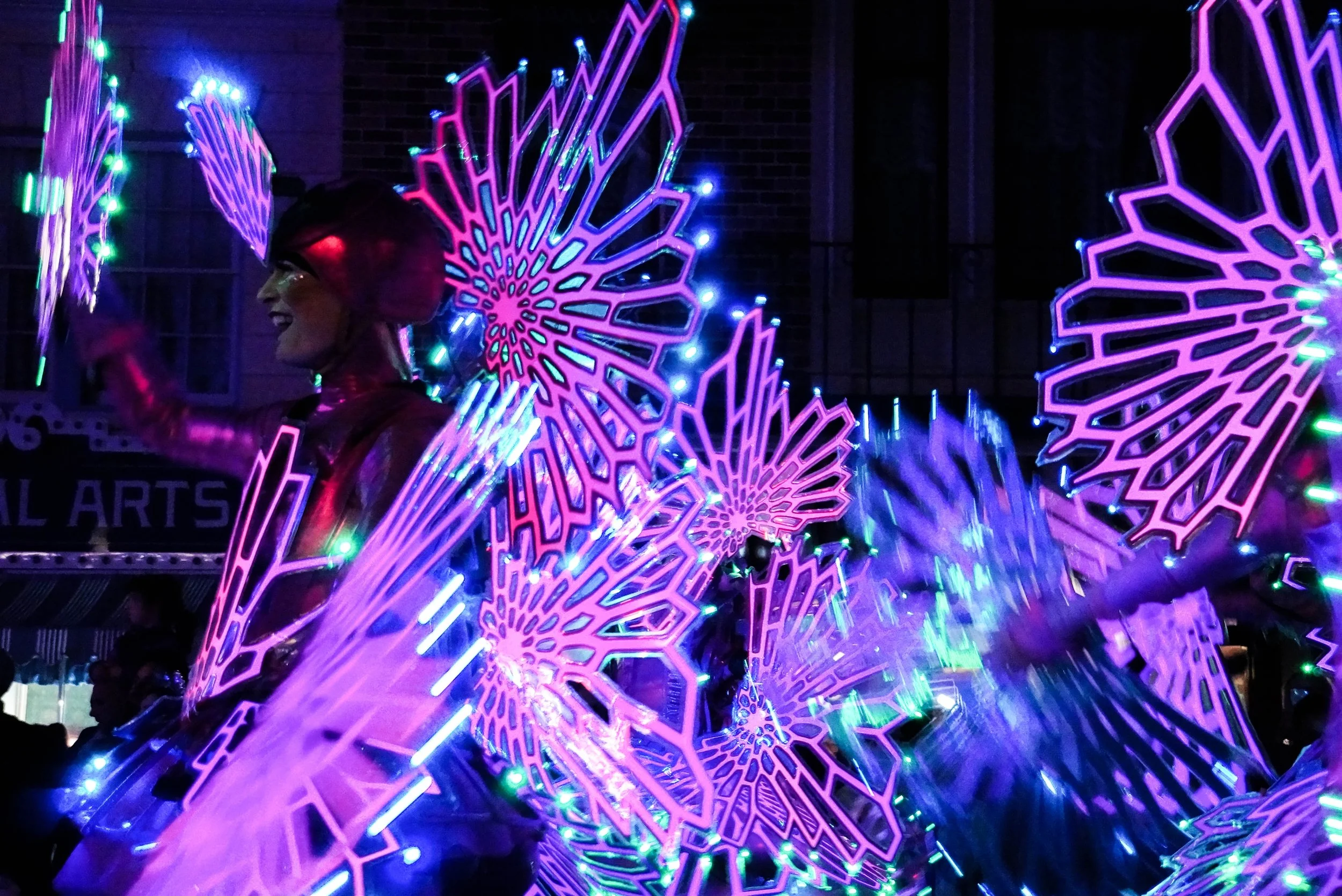 A person wearing illuminated butterfly wings made of pink and purple neon lights at night, smiling and wearing glasses and a shiny pink outfit.