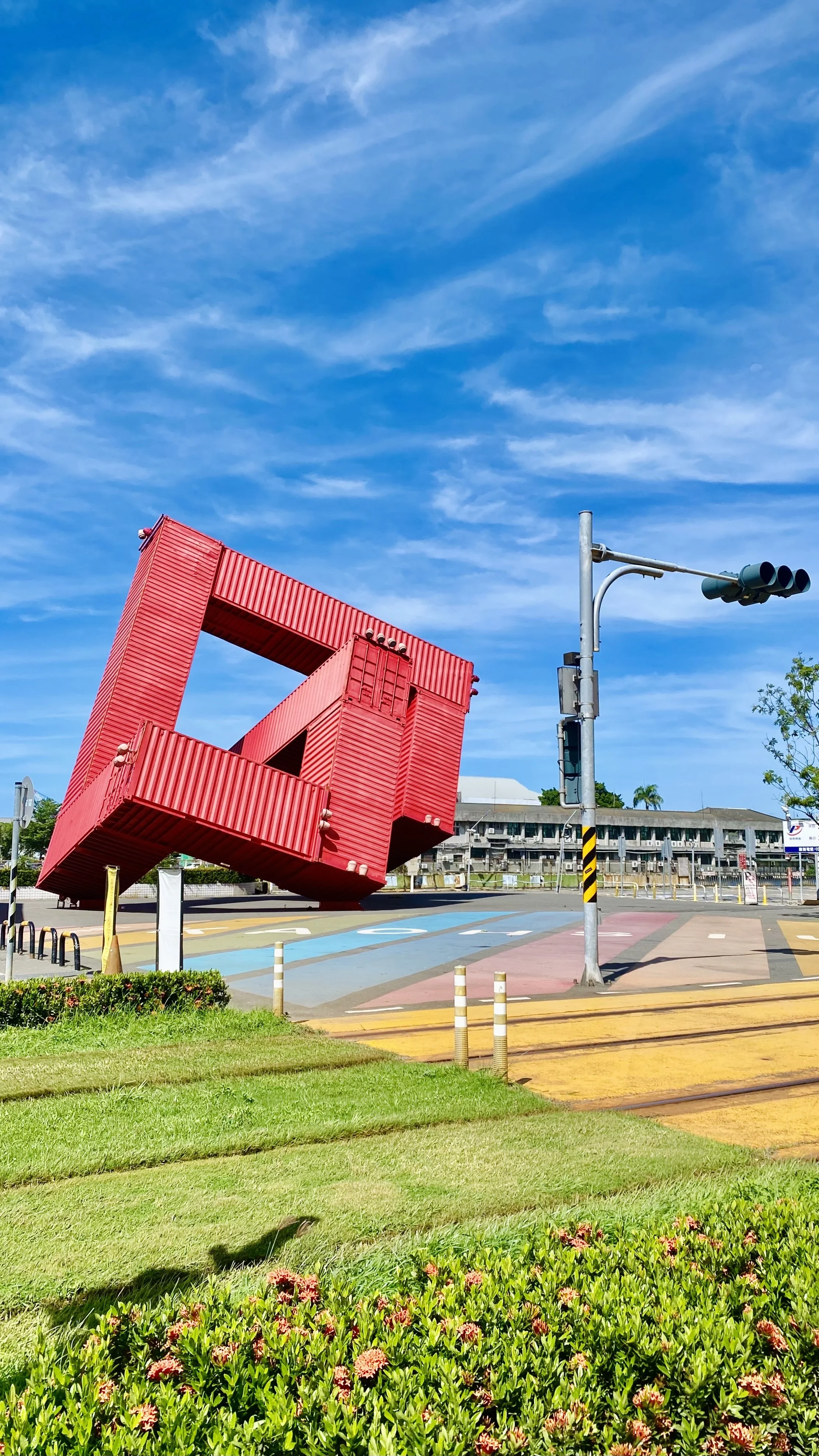 Colorful sculpture made from red shipping containers arranged in a large open loop, set against a bright blue sky with wispy clouds. Traffic lights and a building are visible in the background.
