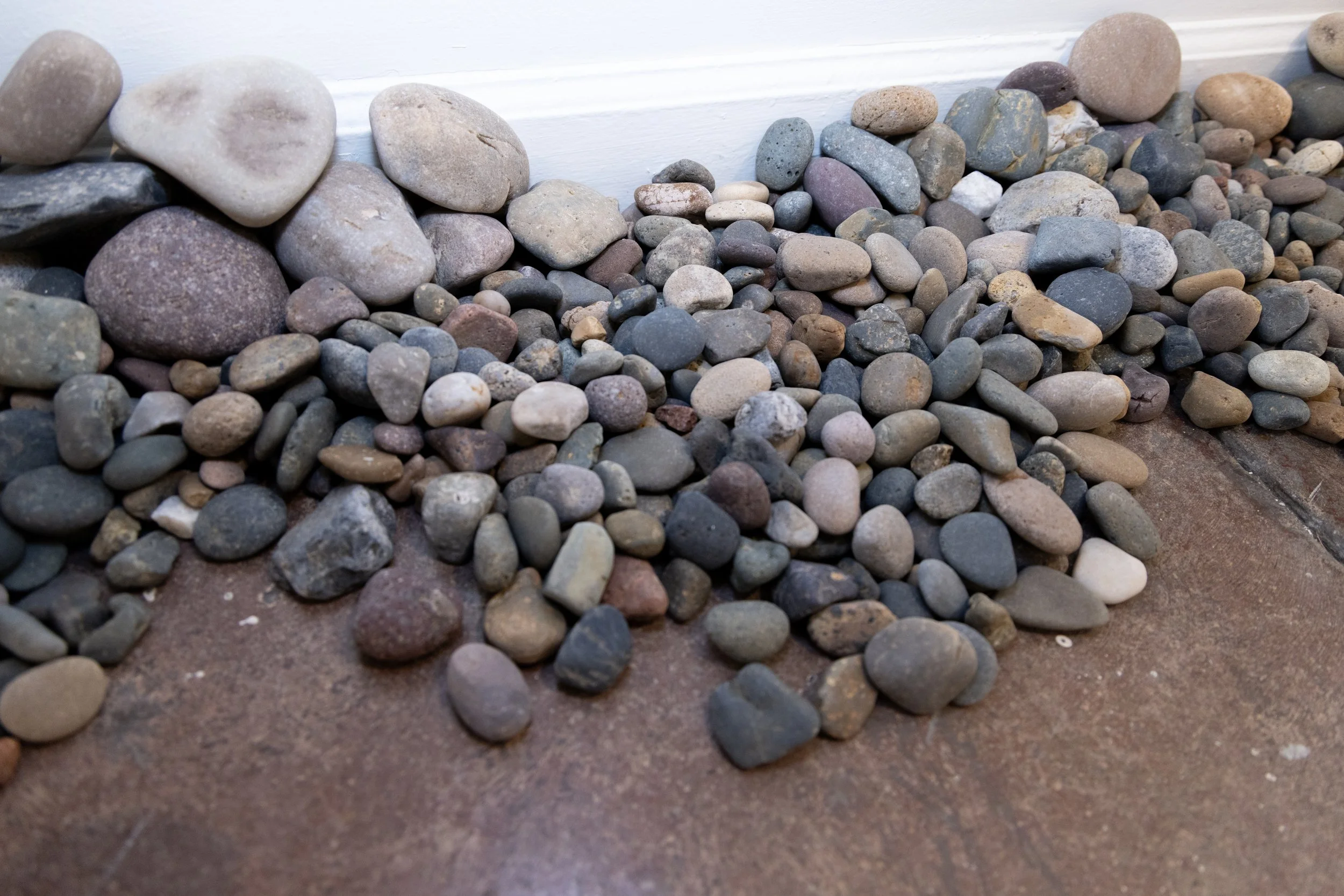 A collection of small to medium-sized colorful smooth rocks and pebbles against a white wall.