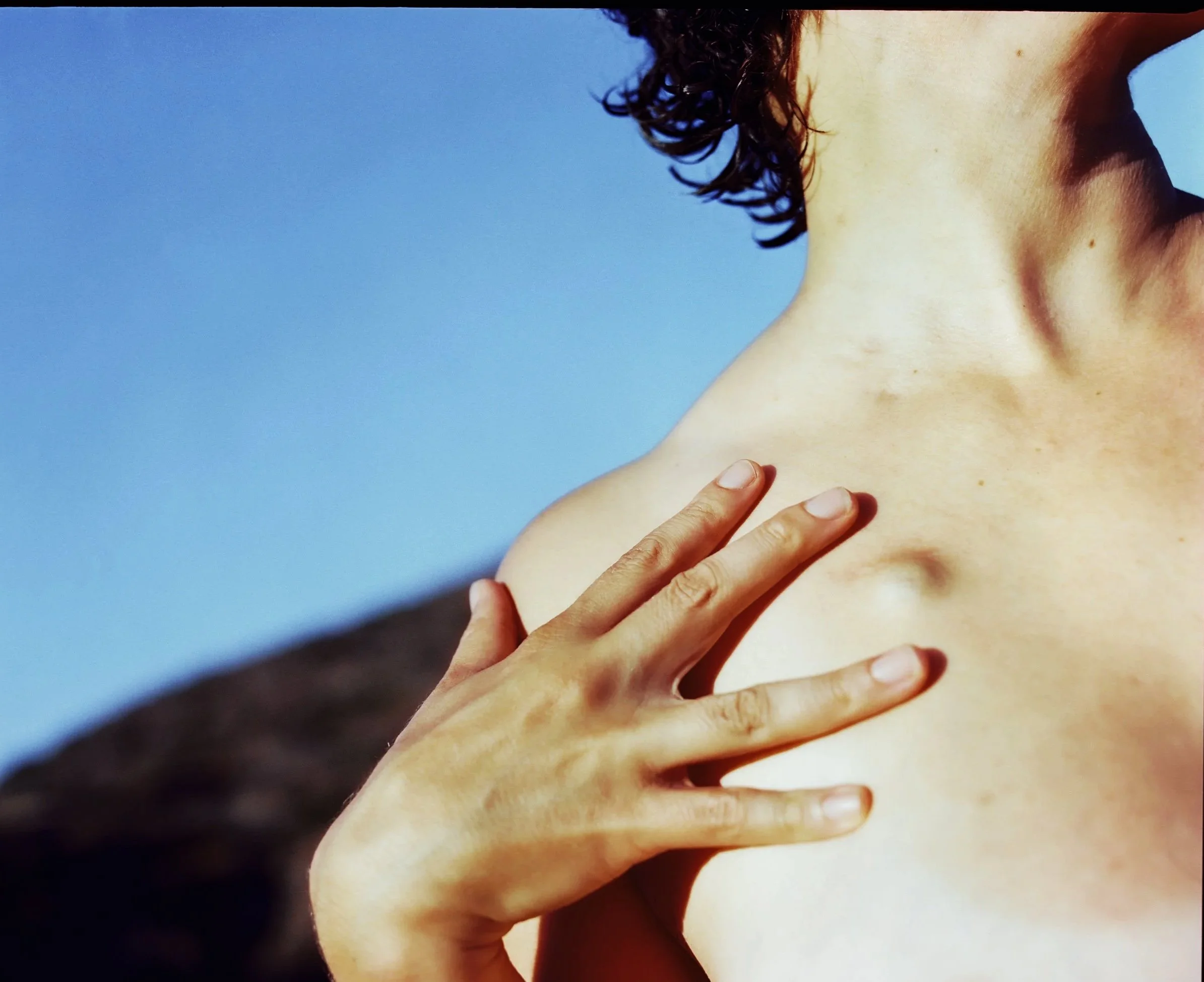 Close-up of a person's hand touching their shoulder and neck against a bright blue sky.
