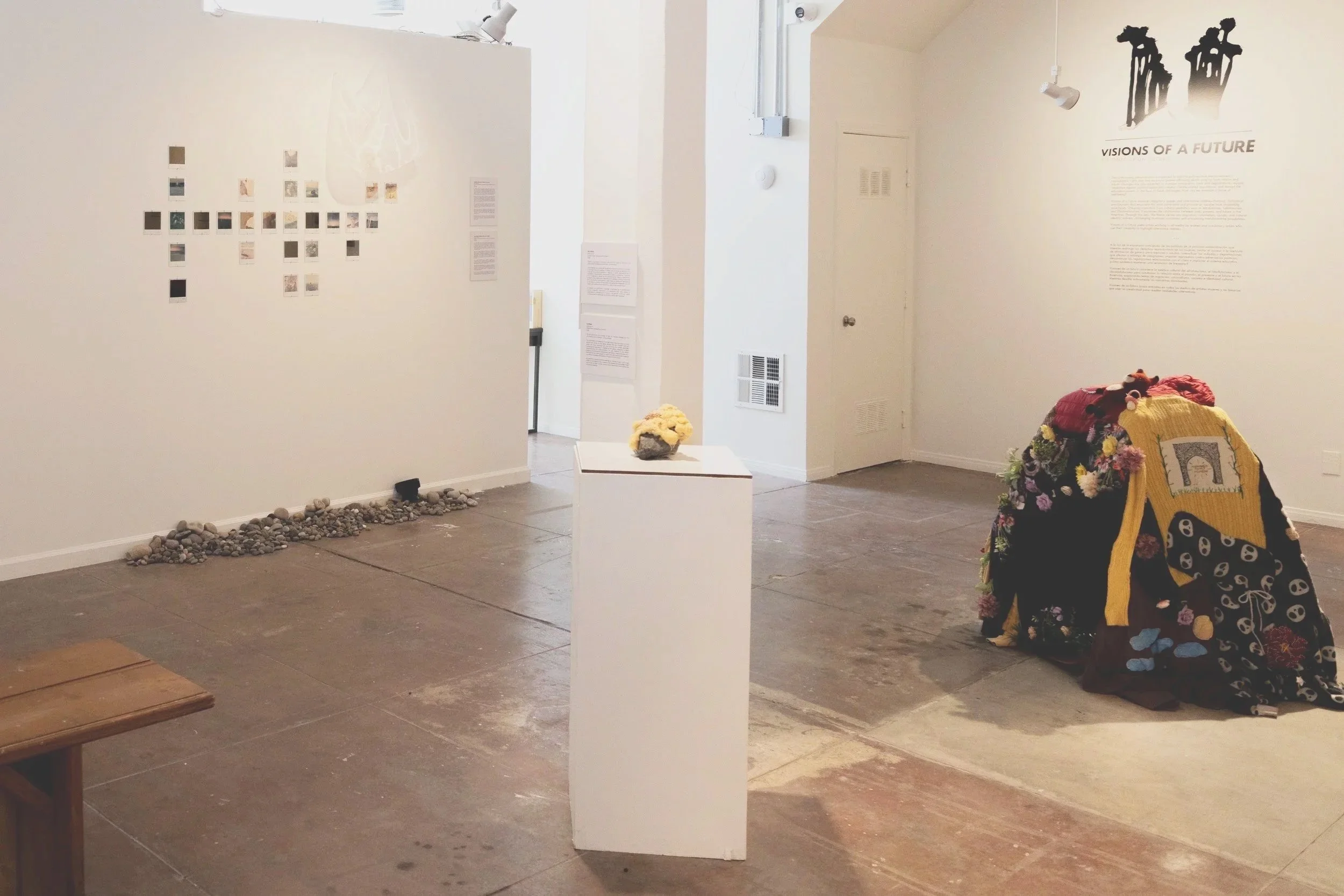 An art gallery displaying an installation titled "Visions of a Future" with black silhouette artwork on the wall. In the foreground, there is a pile of colorful knitted and crocheted items, possibly blankets or sculptures, on the floor and a small wh