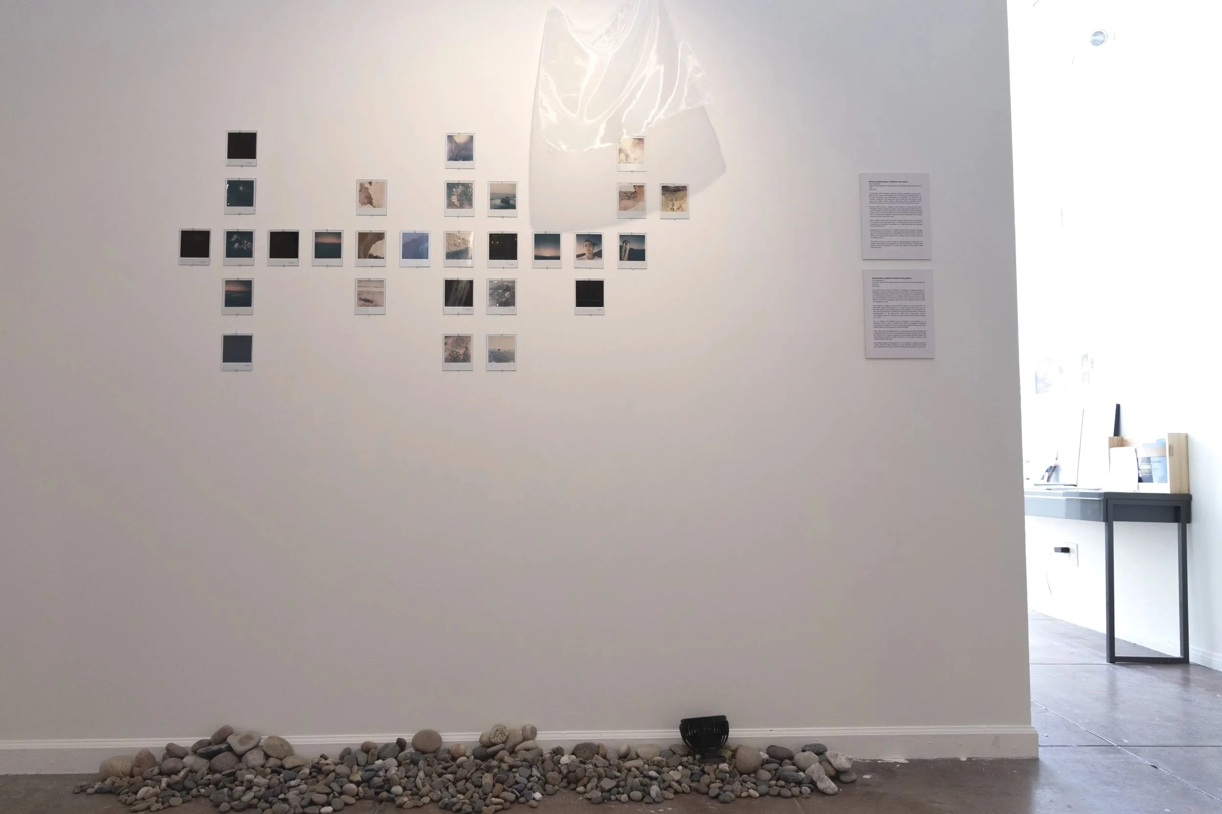 Image of a white gallery wall with small photographs arranged in a semi-circular pattern. Below, there is a collection of rocks on the floor, with a black object and a vent nearby. To the right, an open doorway reveals another room with a counter and