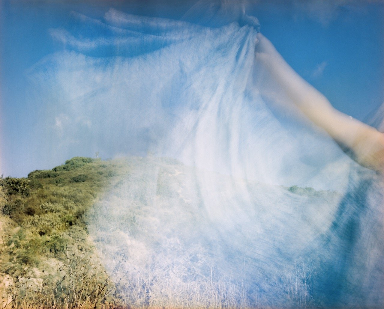 Overlay of a flowing white fabric over a landscape of green hillside under a bright blue sky with wispy clouds.